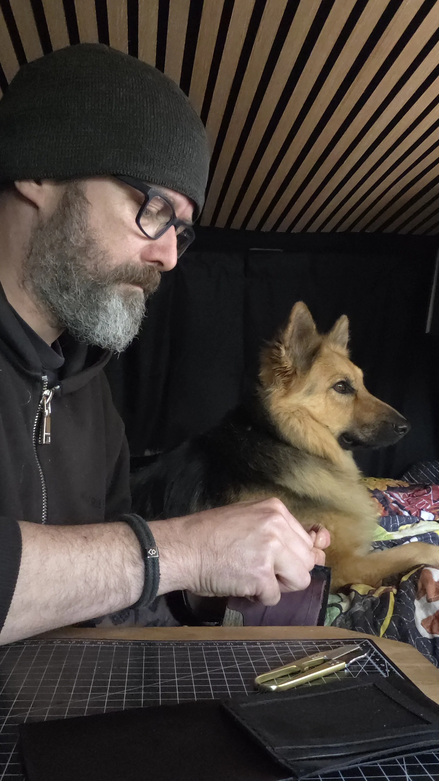 A man with glasses, a black beanie, and a gray beard is sitting at a table with a German Shepherd dog. The man is holding the dog's paws, and they are both looking to the right. There are cutting tools and a grid cutting mat on the table.