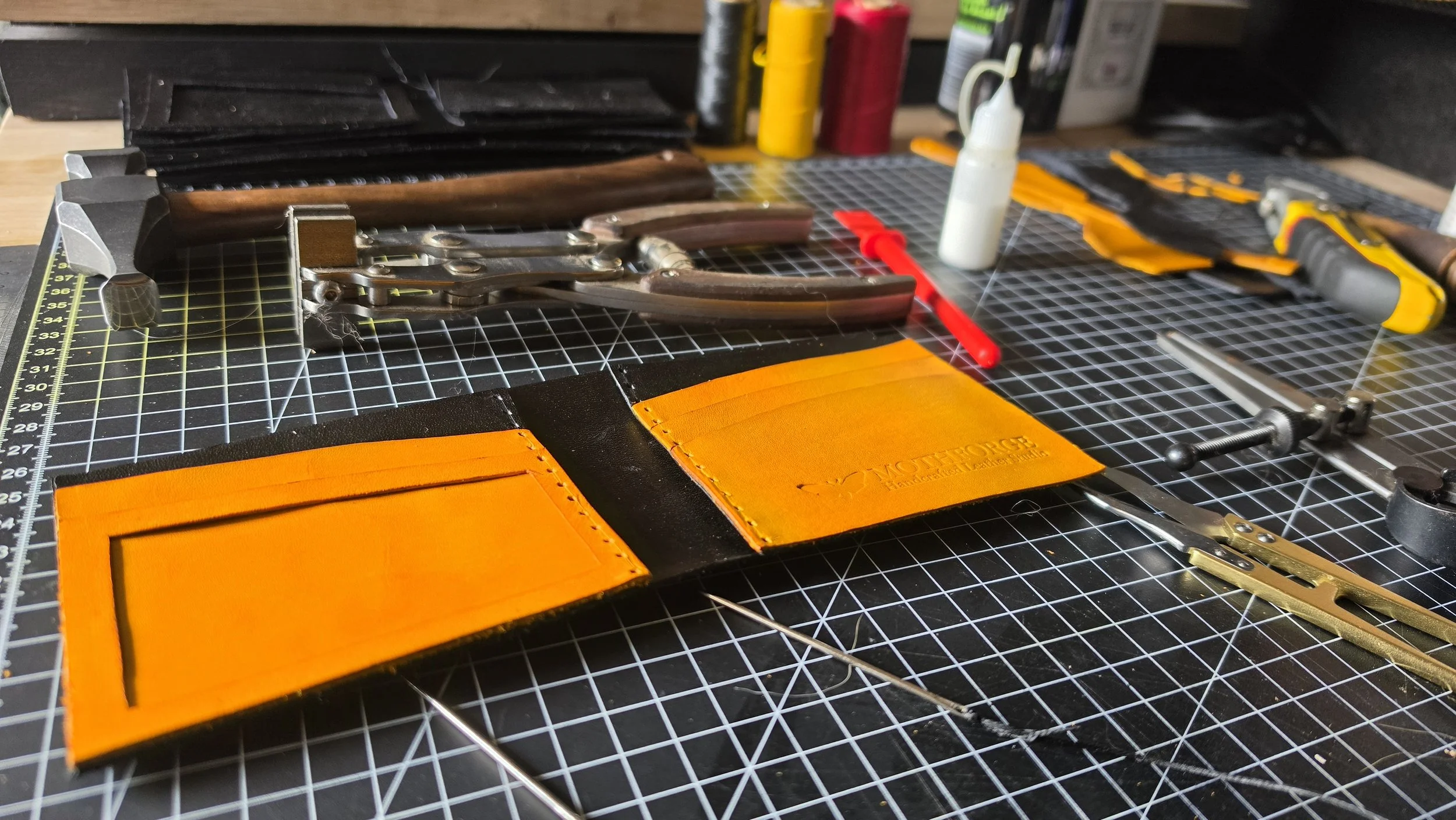 Workstation with leatherworking tools including scissors, knives, and glue, along with orange leather pieces on a cutting mat.