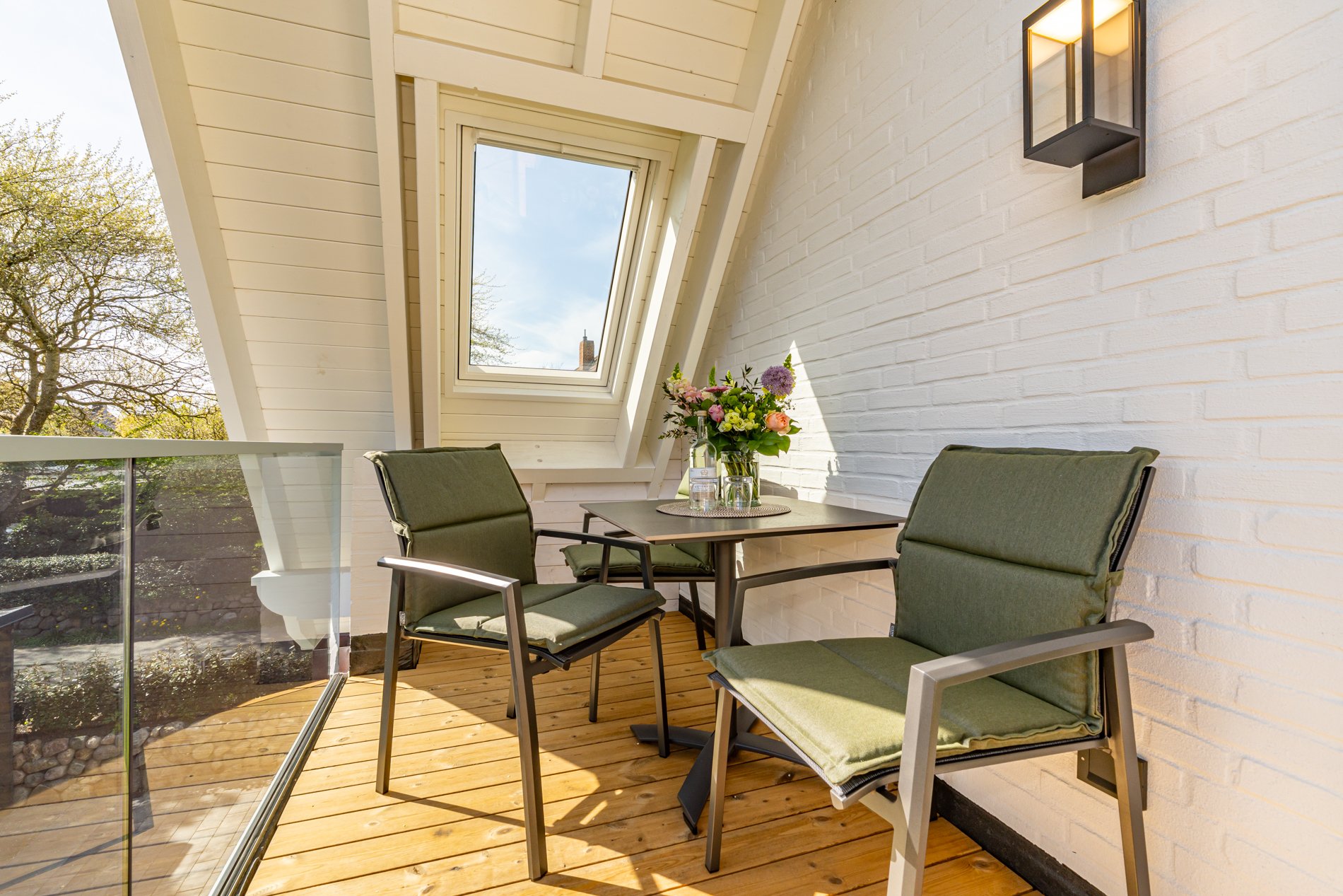 Ein Balkon mit Holzboden, einer weißen Backsteinwand, einem schwarzen Wandleuchter, einem Tisch mit einer Blumenampel, zwei grünen Stühlen und einem großen Dachfenster, durch das der Himmel und einige Bäume sichtbar sind.