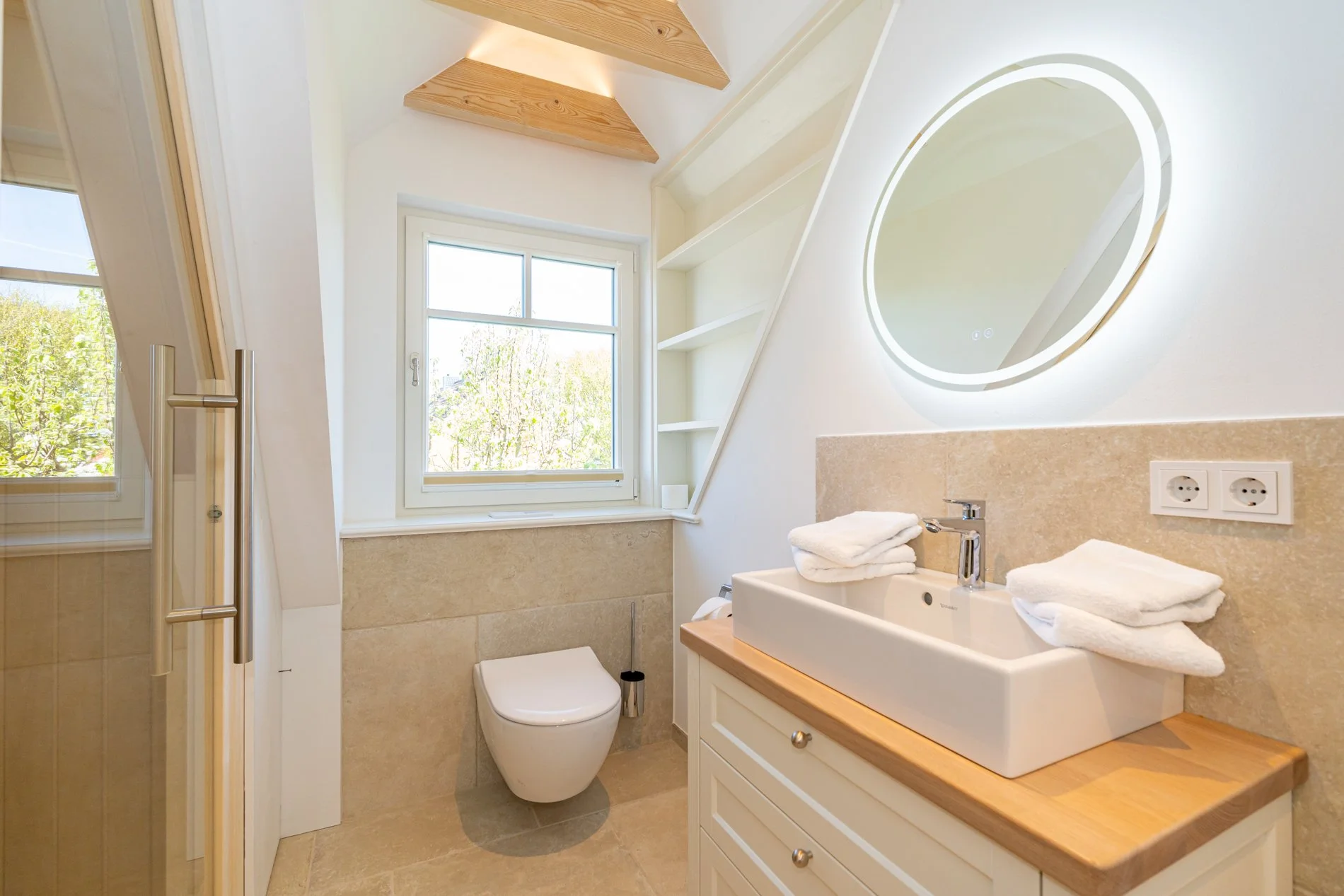 Ein helles Badezimmer mit einem grossen runden Wandspiegel, Waschbecken, Handtüchern, Fenster, Toilette und einer Dusche
