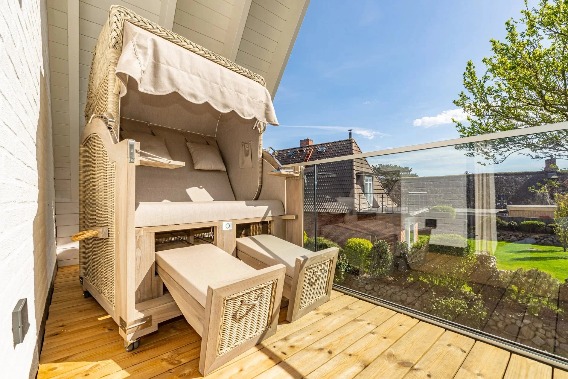 Ein gemütlicher Balkon mit Holzfußboden, ausgestattet mit einem geflochtenen Gartensessel mit Dach, einem kleinen Tisch und verglasten Geländern, mit Blick auf ein Nachbarhaus und einen blauen Himmel.