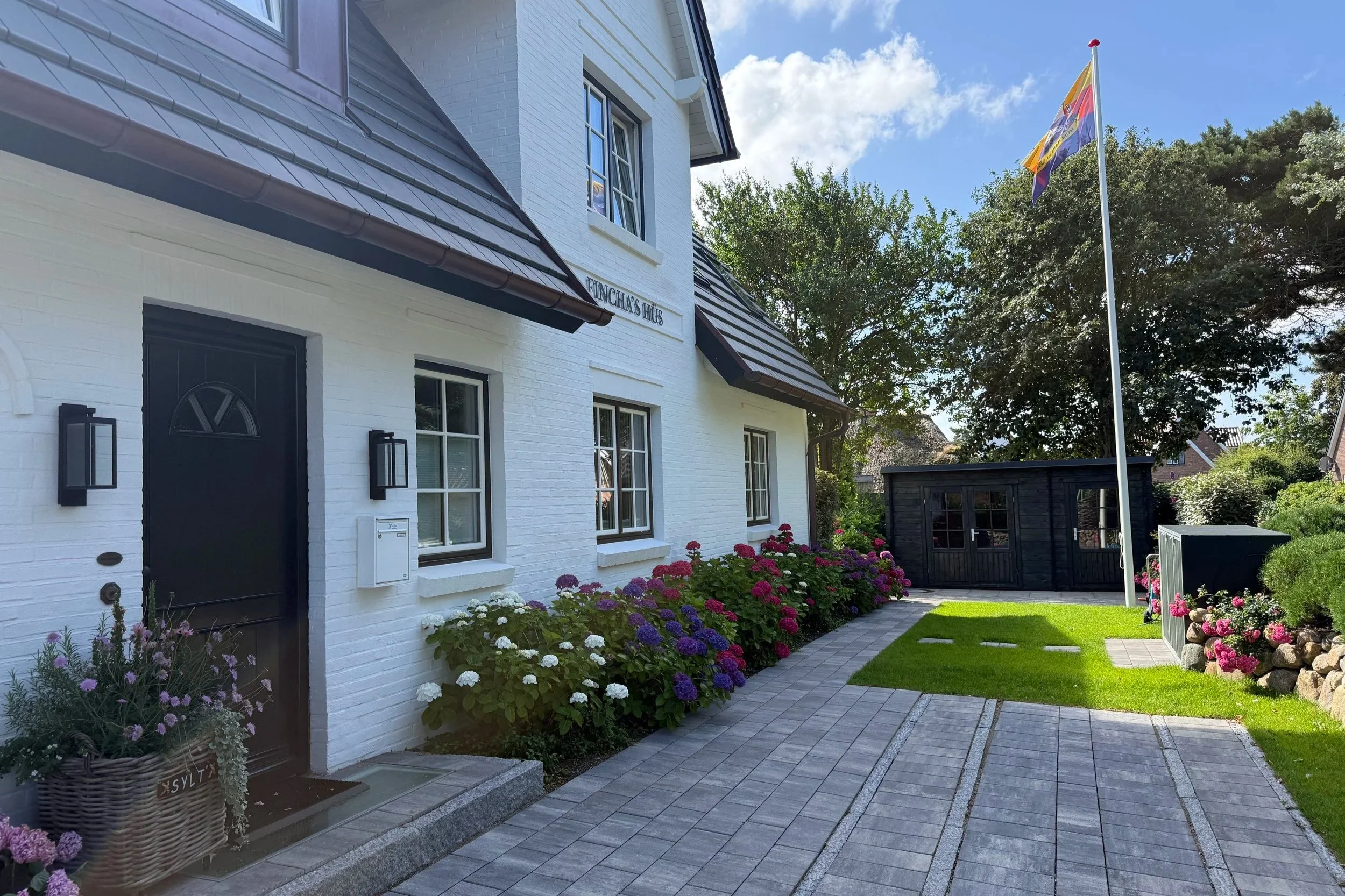 Ein weißes Haus mit schwarzer Tür, umgeben von bunten Blumen und einer gepflegten Rasenfläche. Es gibt einen gepflasterten Weg und einen Garten mit Bäumen und einem schwarzen Gartenhaus. Eine Flagge weht auf einem hohen Flaggenmast.