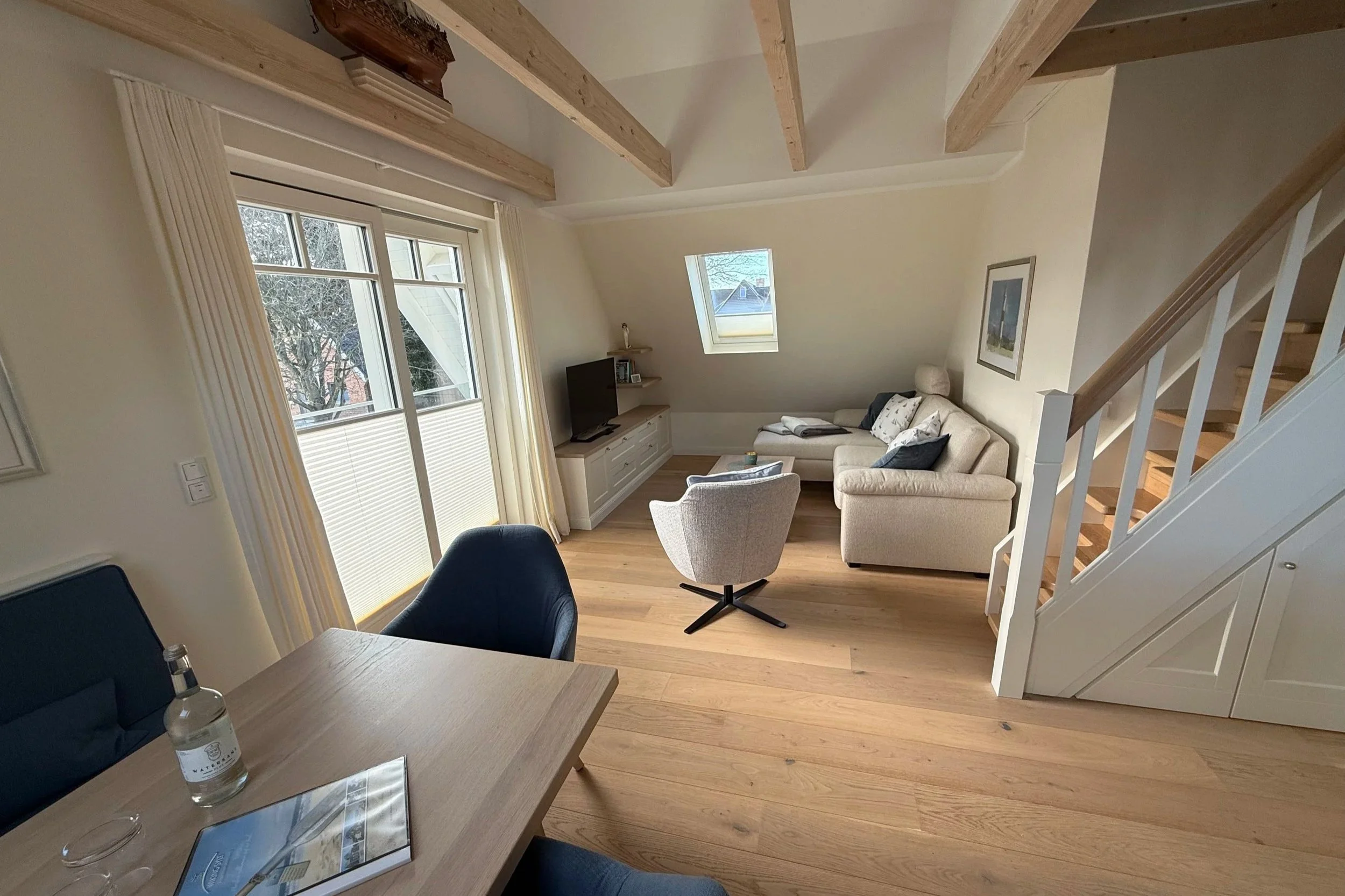 Innenaufnahme eines Wohnzimmers mit großen Fenstern, Couch, Fernseher, Stuhl und Holzboden sowie sichtbaren Deckenbalken.