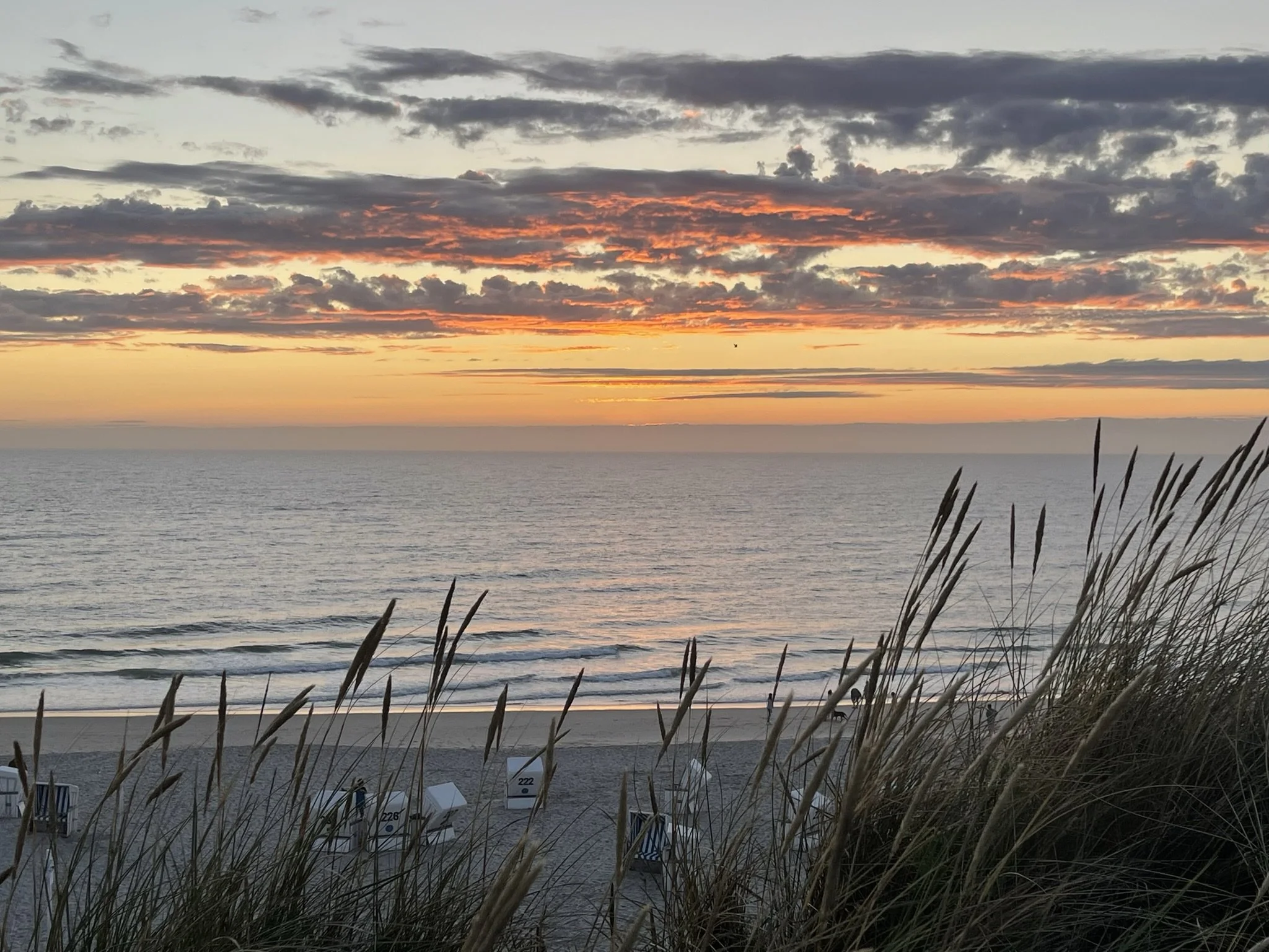 Typischer Nordsee-Strand mit Strandkörben, im Hintergrund der Ozean und ein Sonnenuntergang mit buntem Himmel und Wolken.