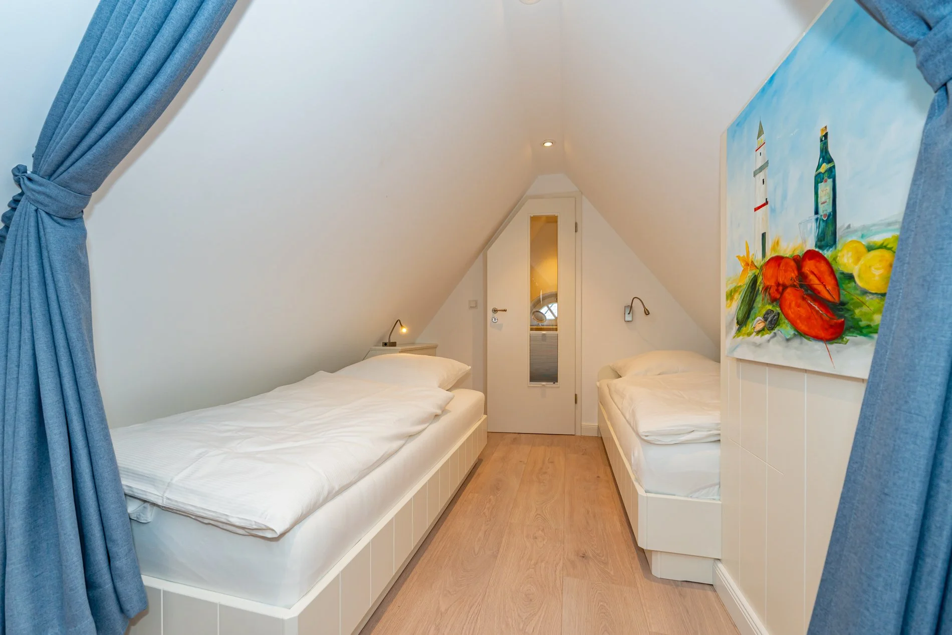Ein kleines Schlafzimmer mit zwei Betten unter einer Dachschräge, blau gestrichenen Vorhängen, einer bunten Gemälde an der Wand, weißen Möbeln und Holzboden.