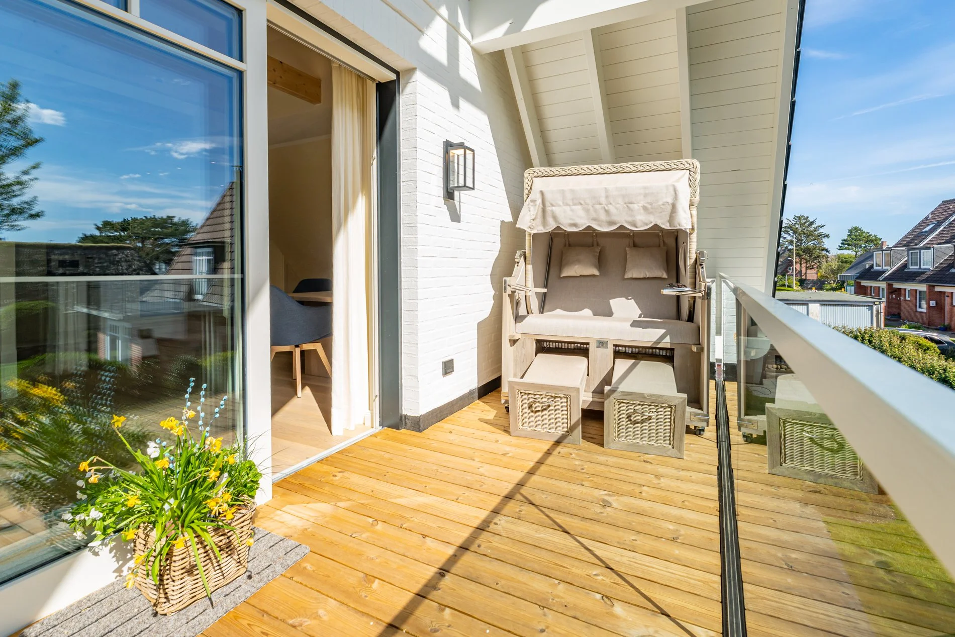 Ein sonniger Balkon mit Holzdielen, ein Strandkorb, grüne Pflanzen in einem Korb, und ein Blick auf Nachbarhäuser mit grünen Gärten.