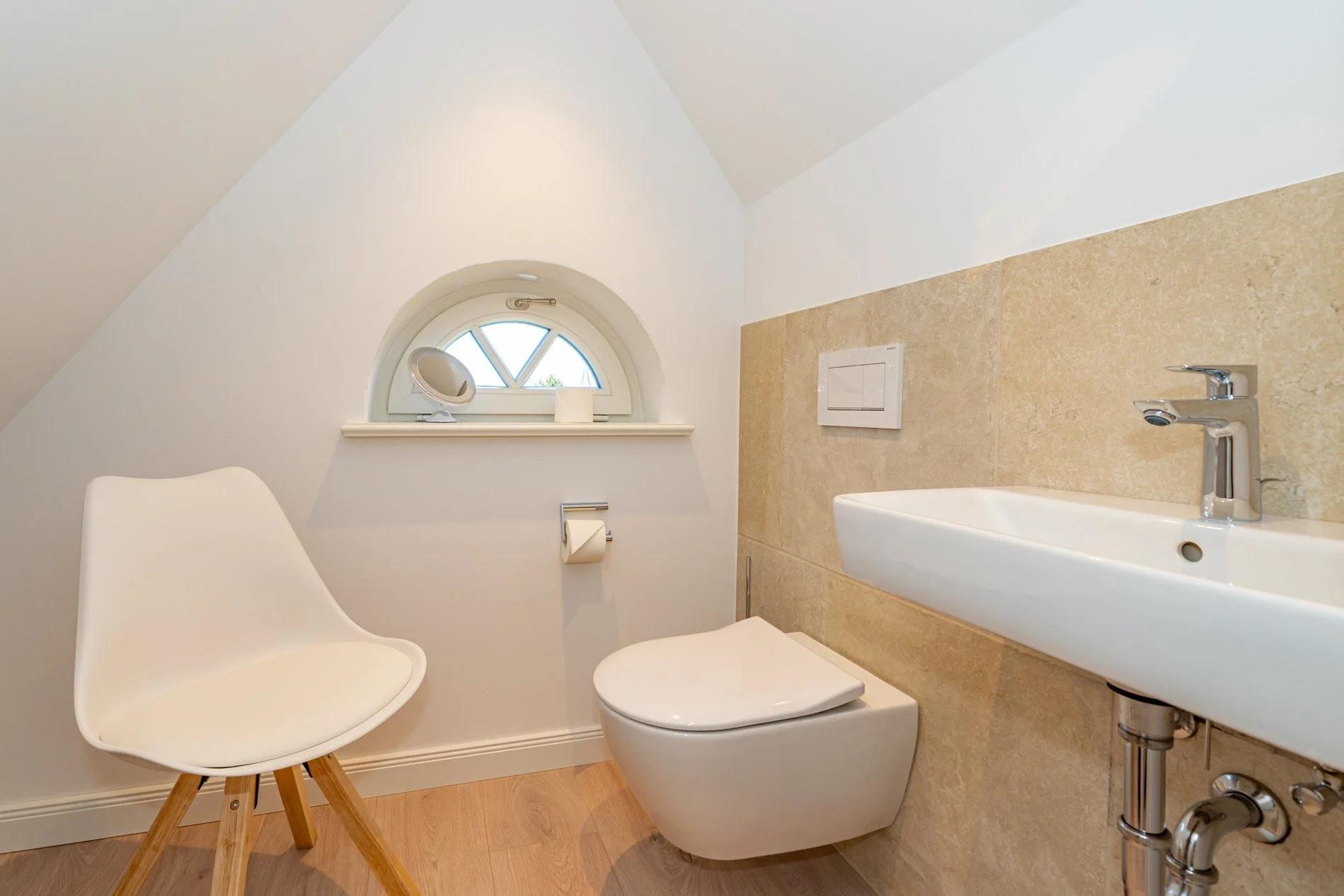 Ein kleines Badezimmer mit weißem WC und einem weißen Waschbecken, einem weißen Stuhl, einer kleinen runden Fensterbank mit Spiegel, Toilettenpapierrolle, und einem Fenster mit arched Oberteil.