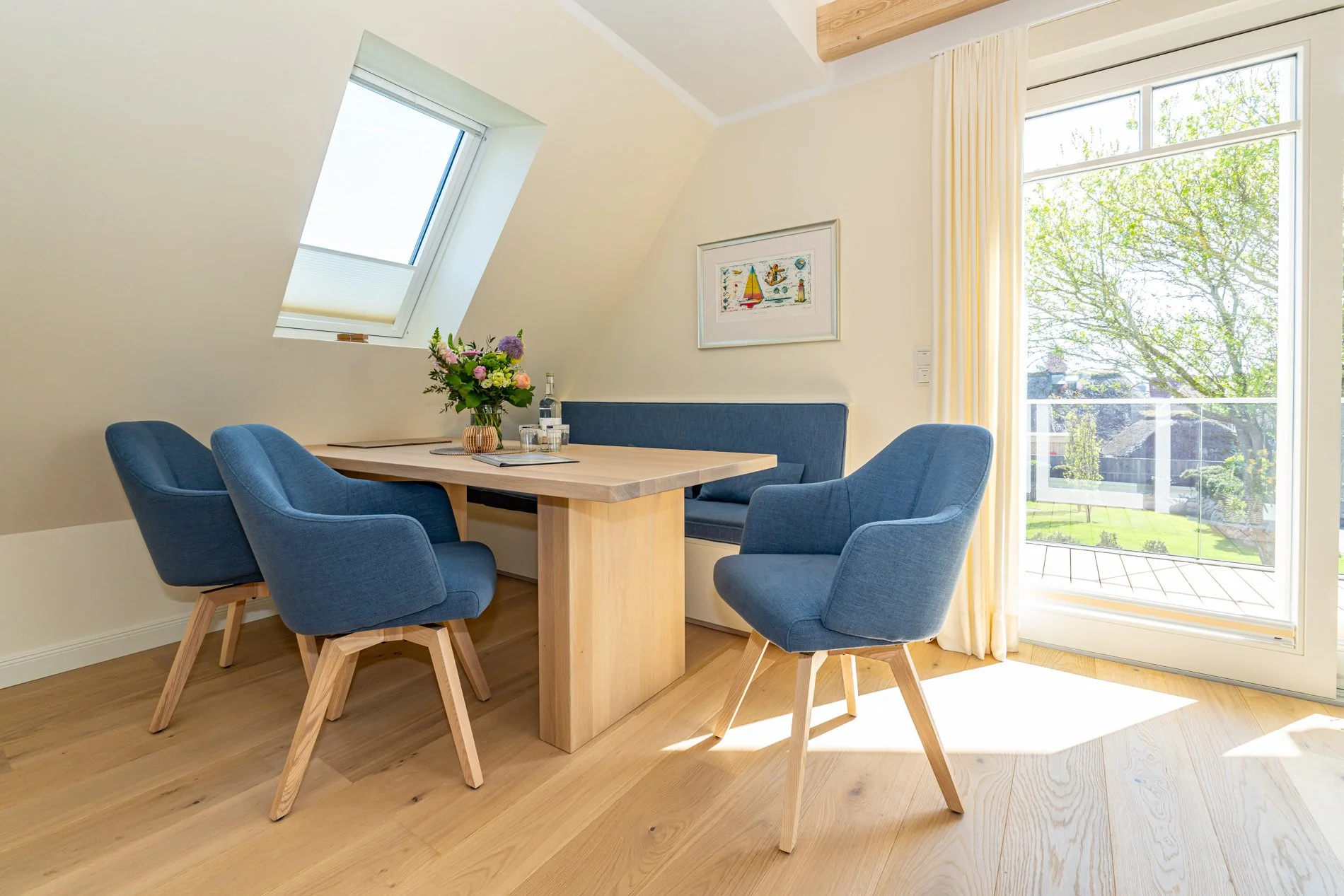 Ein heller Raum mit einem Holzfußboden. Es gibt einen runden Tisch mit vier blauen Sesseln. Im Raum hängen gelbe Vorhänge, und es gibt große Fenster, die nach draußen auf einen Garten mit Bäumen blicken. An der Wand hängt ein Bild.