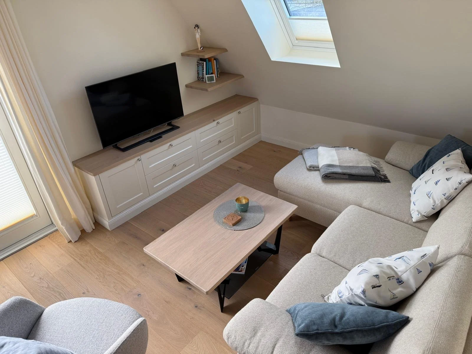 Ein gemütliches Wohnzimmer mit einer Couch, einem Couchtisch, einem Flatscreen TV auf einem weißen Lowboard, einem schrägen Dachfenster und einer kleinen Ecke mit Bücherregale.