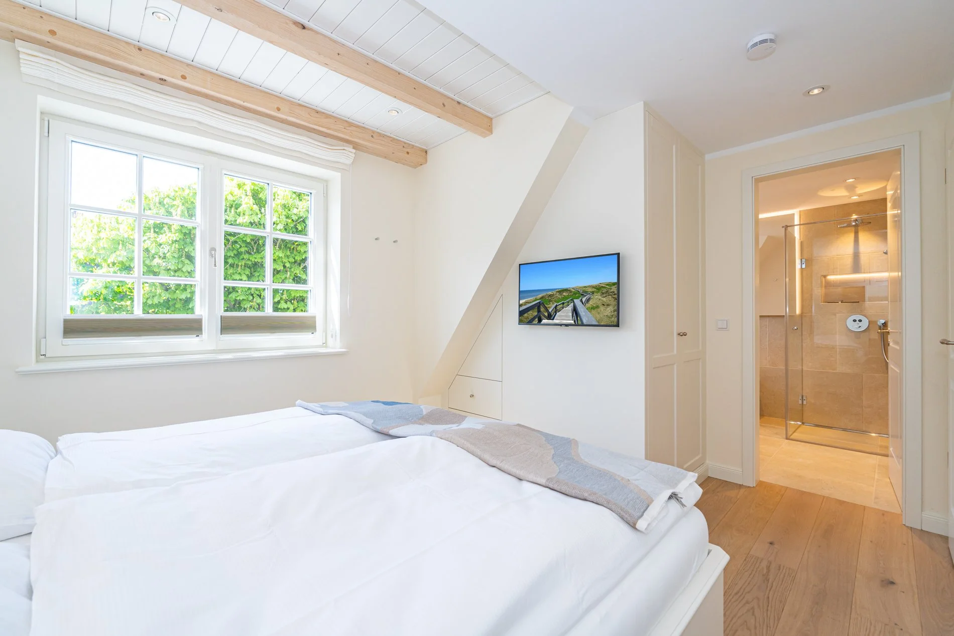 Ein Schlafzimmer mit weißen Betten vor einem Fenster, das grünen Baum außerhalb zeigt, und einem Fernseher an der Wand. Im Hintergrund ein Badezimmer mit begehbarer Dusche.