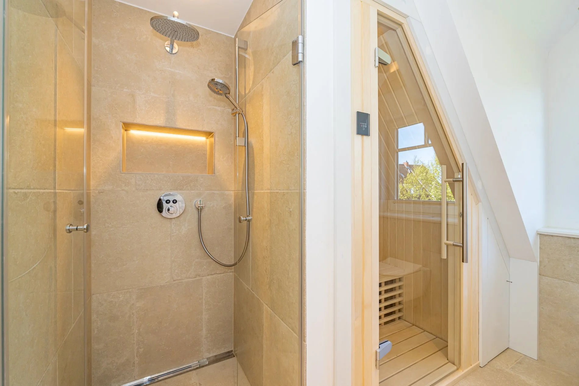 Duschbereich mit zwei Duschköpfen und einer Nische, daneben eine Sauna in Holzoptik mit Glasfront und Temperaturschalter, mit sichtbarem Blick nach draußen auf Bäume und Himmel.