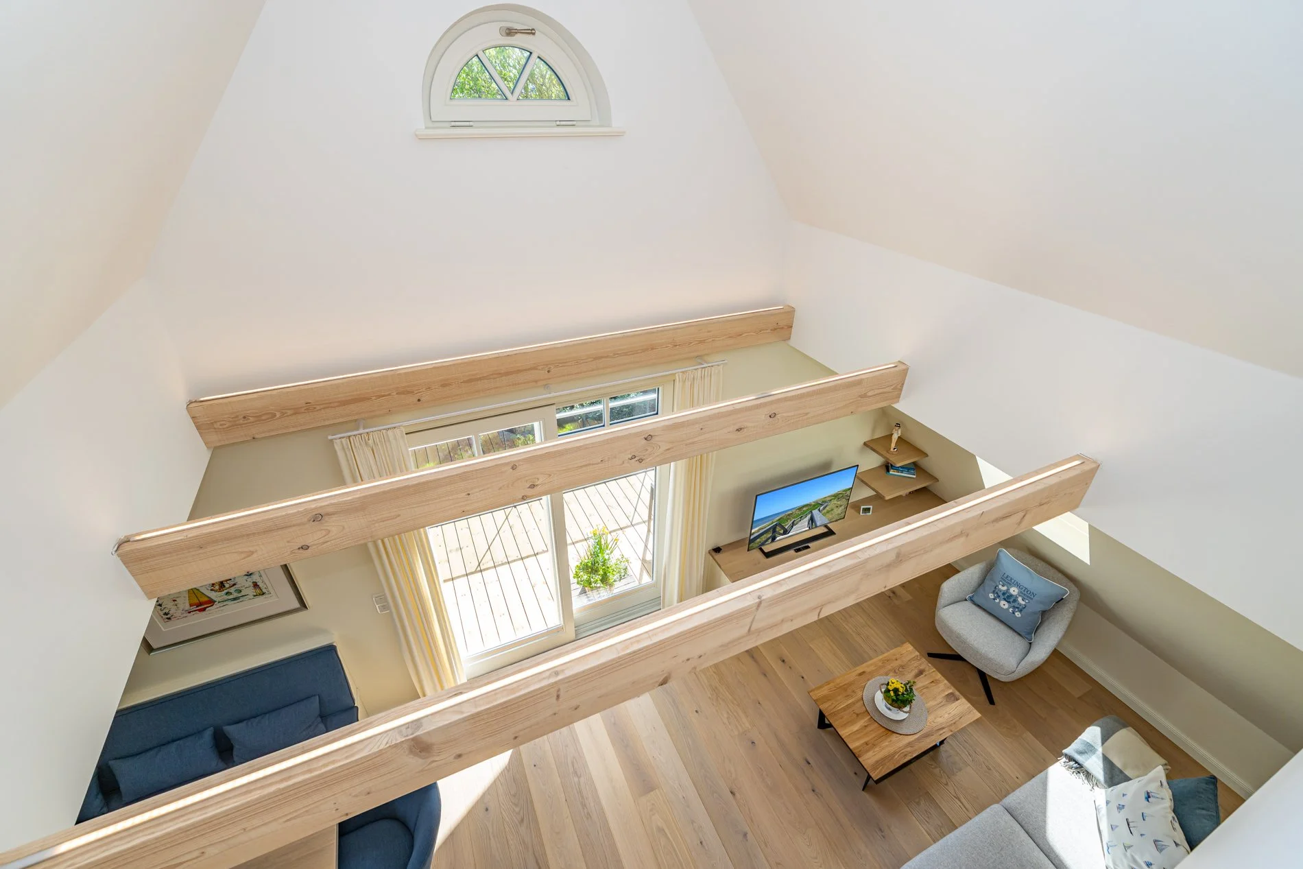 Innenaufnahme eines mehrstöckigen Wohnzimmers mit Holzbalken (Perspektive von oben), großen Fenstern und moderner Einrichtung, darunter Sofas, Fernseher und Türen nach draußen.