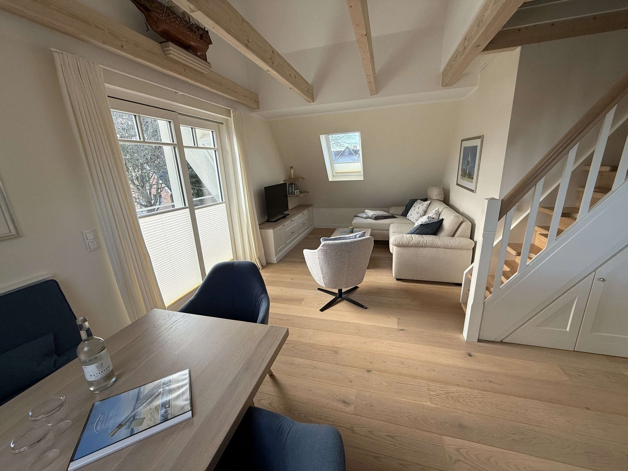 Wohnzimmer mit Holzdeckenbalken, großen Fenstern, einem Fernseher auf einem Medienmöbel, Sofa, Sessel, und Treppe im Hintergrund.