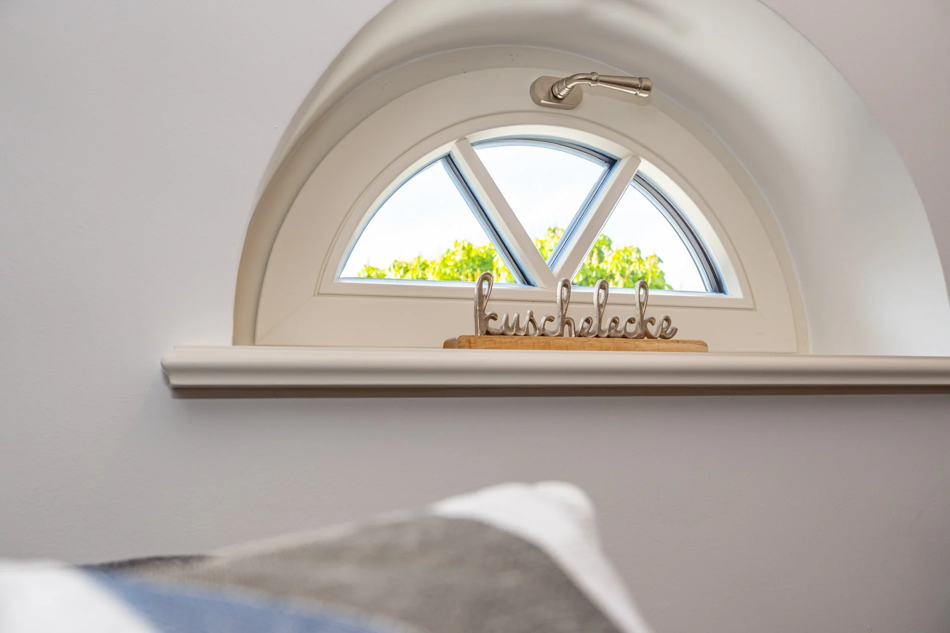 Kleines halbkreisförmiges Fenster mit Blick nach draußen, durch das man grüne Bäume und Himmel sieht.