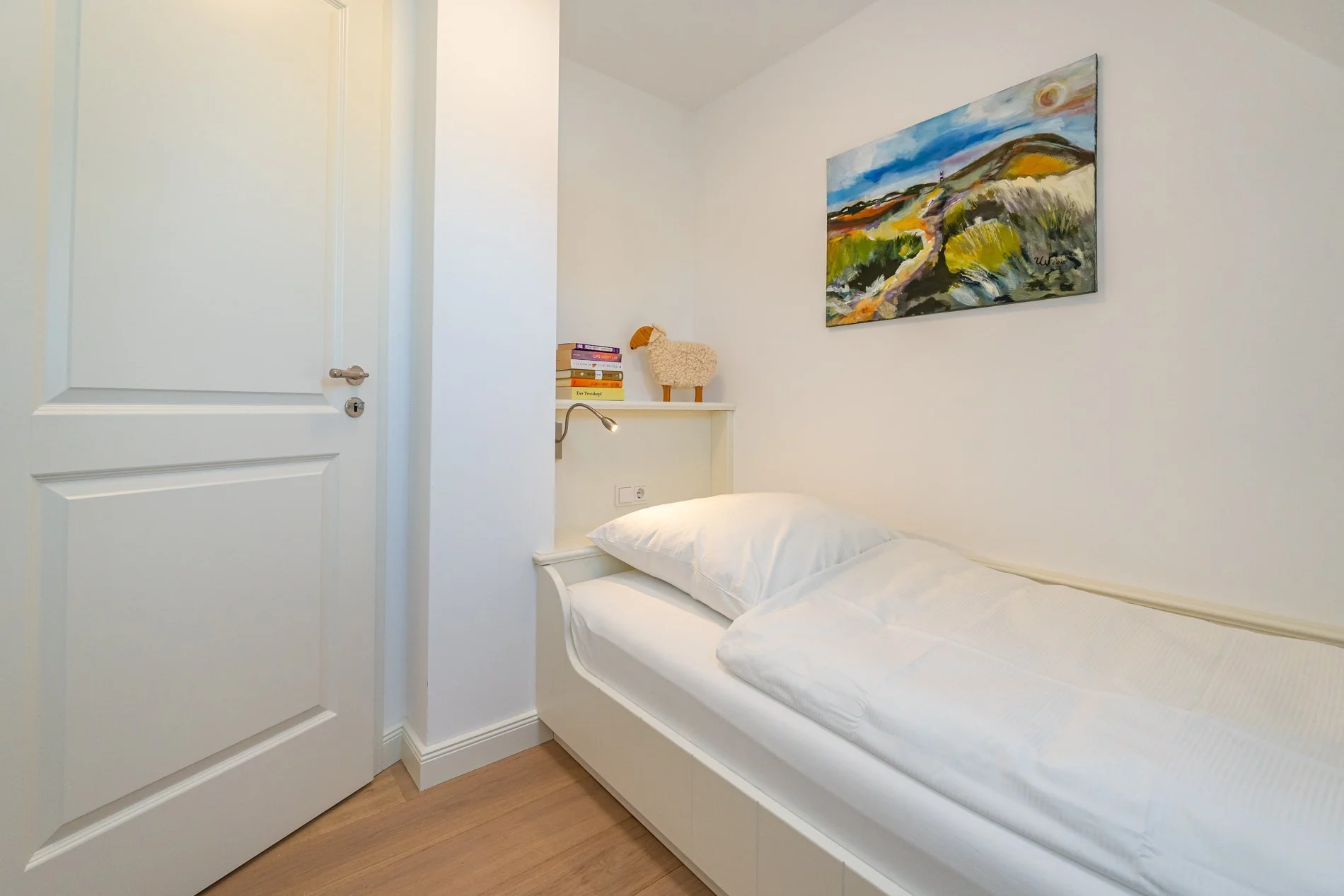 Ein Schlafzimmer mit weißem Bett, einer weißen Wand und einem bunten, abstrakten Landschaftsbild an der Wand. Es gibt eine weiße Tür und eine kleine Nachttischlampe neben dem Bett, sowie einige Bücher und ein dekoratives Schaf auf einem Regal.