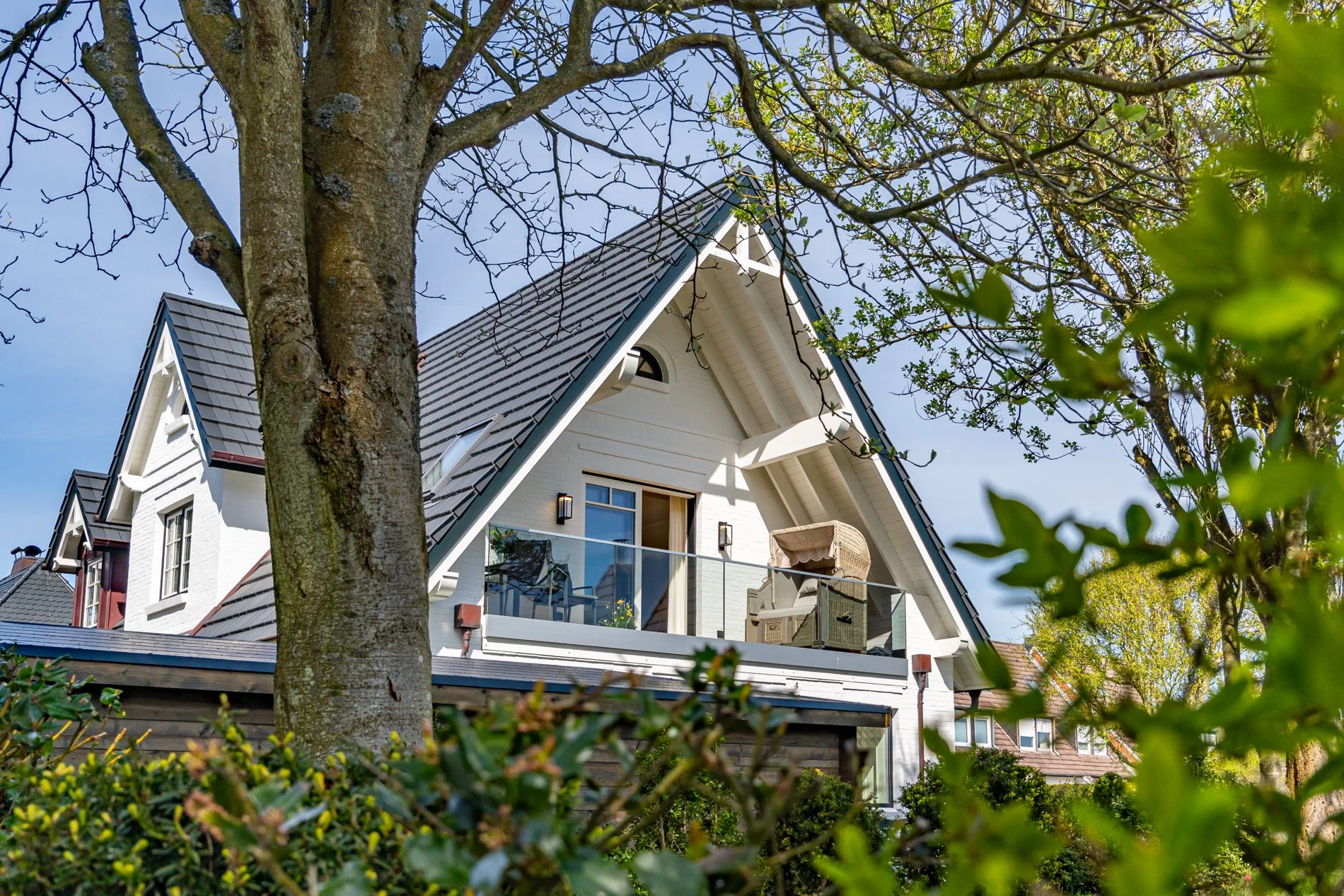 Ein modernisiertes Haus mit weißer Fassadenfarbe, großen Fenstern und einem Balkon, teilweise hinter Büschen und Bäumen. Der Blick ist nach oben, mit blauen Himmel im Hintergrund.