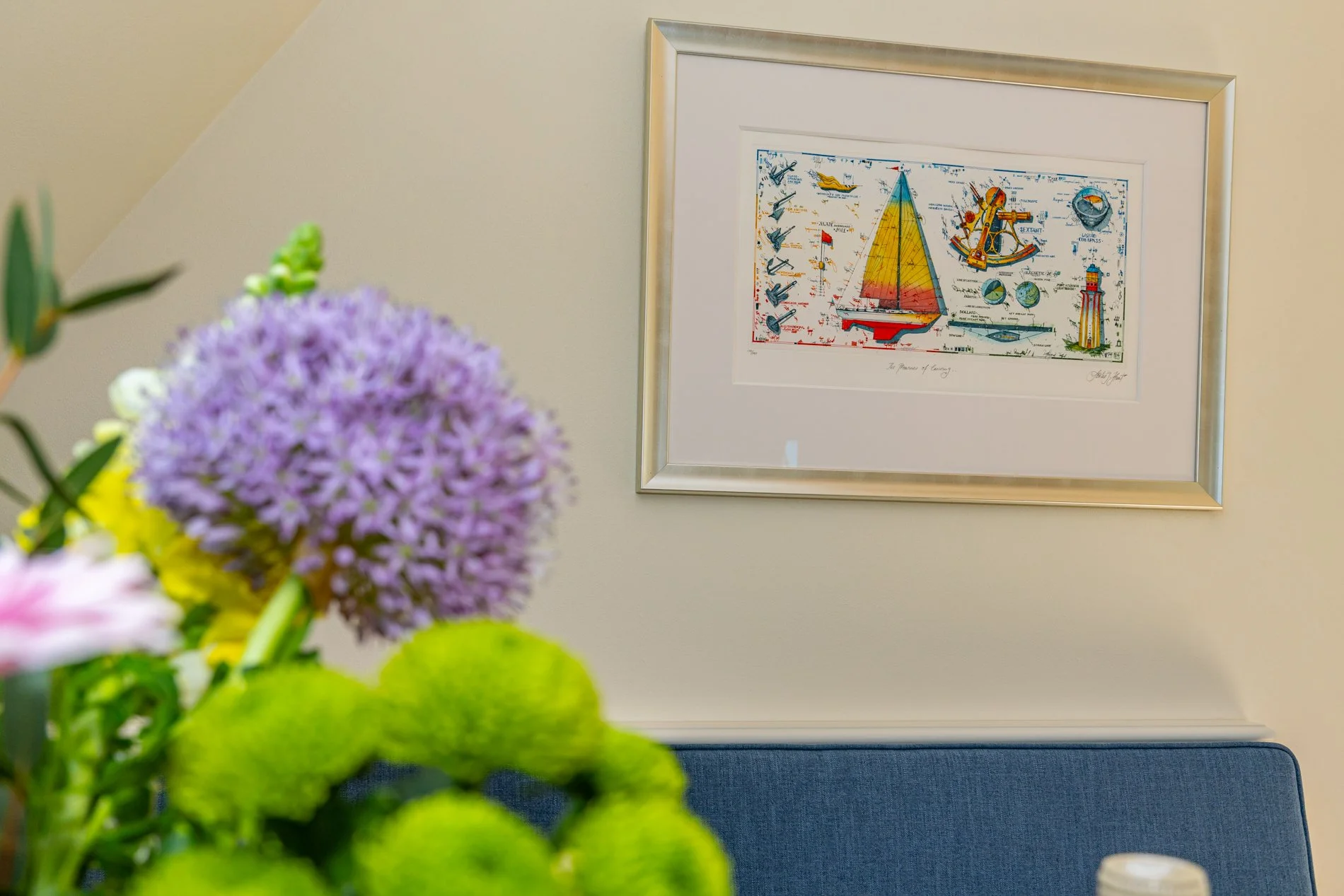Bunte Blumen im Vordergrund, im Hintergrund ein gerahmtes Seefahrtbild an der weißen Wand.