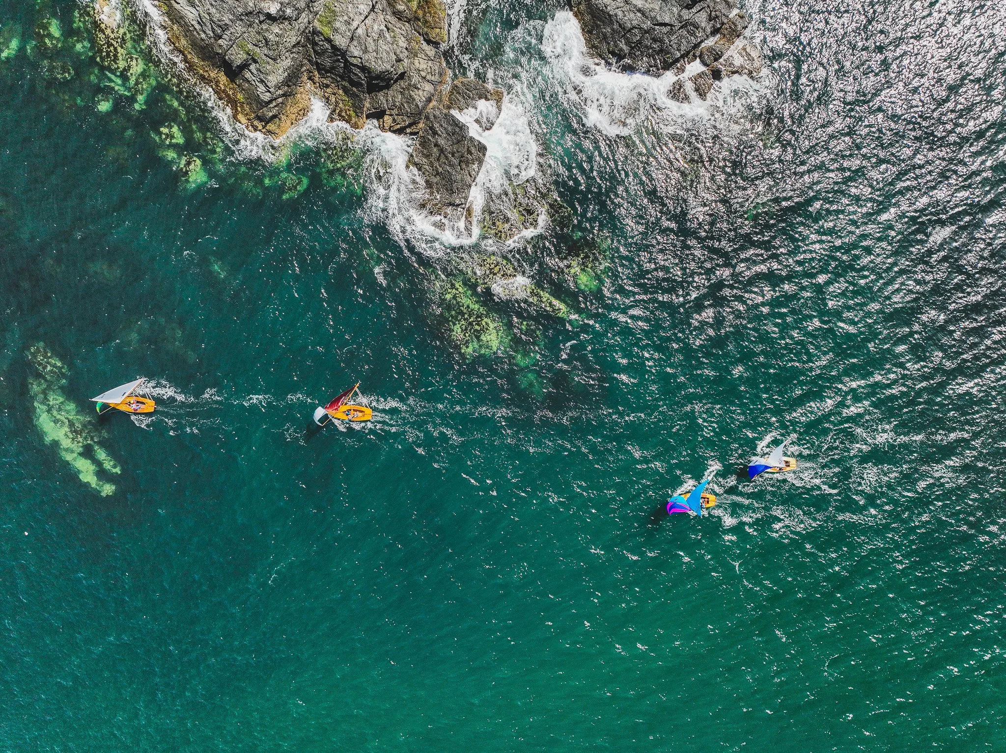 Aerial view of five small sailboats with colorful sails sailing near rocky coastline with waves crashing against rocks.