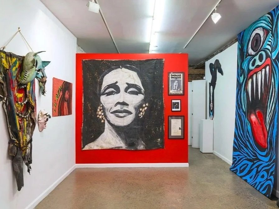Art gallery with a red wall displaying a large painting of a woman's face, surrounded by various abstract and expressive artworks, including a colorful dragon mask on the right and a creature mask with horns on the left.