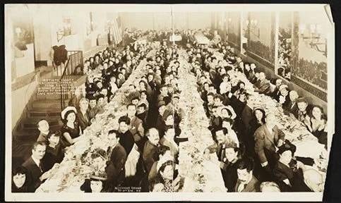 A large group of women and children seated at long dinner tables in a large hall, possibly a banquet or community gathering in the early 20th century.