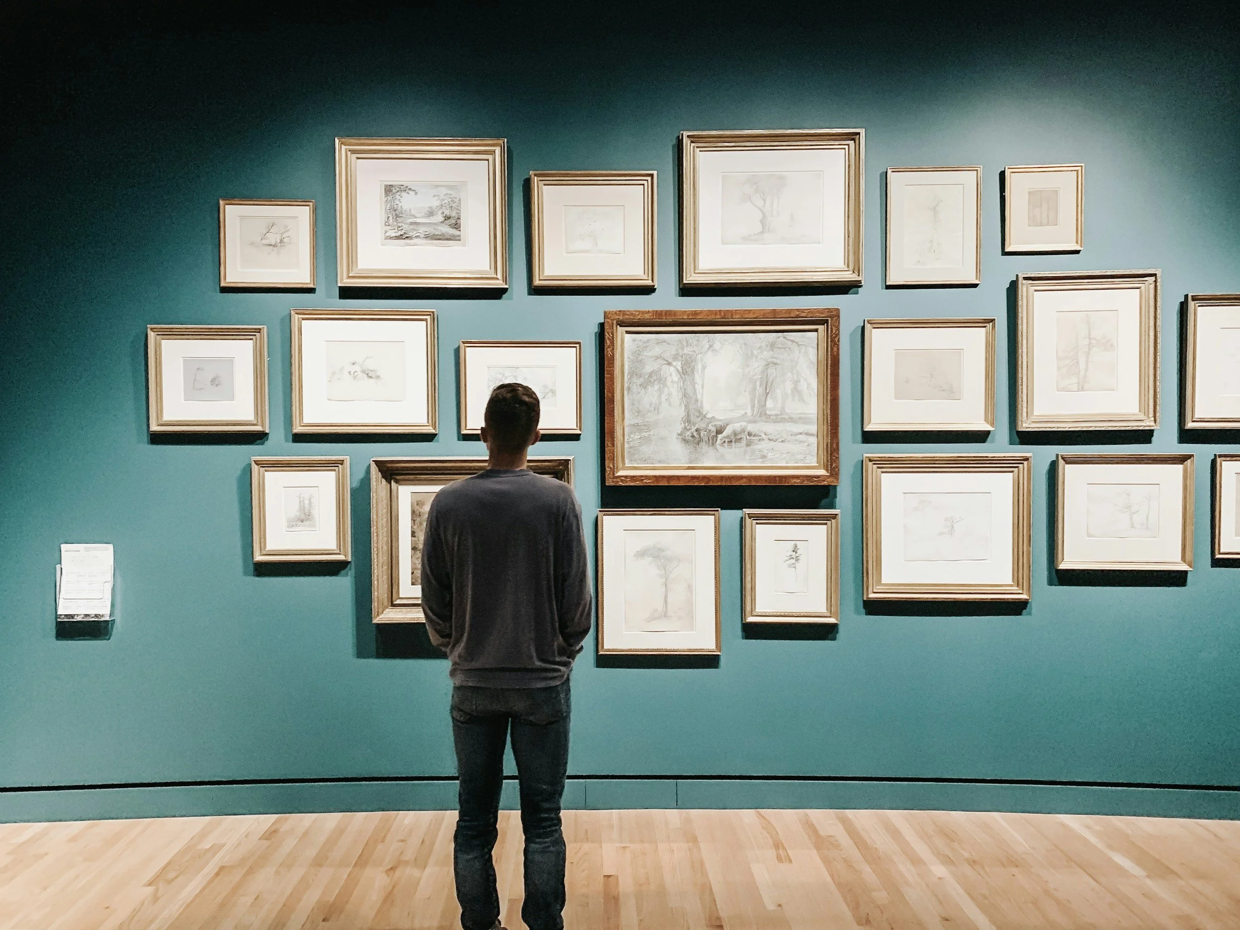 A man stands in front of a green wall with multiple framed artworks in a museum or gallery, observing the art.
