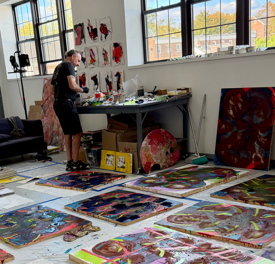 An artist working in a bright studio surrounded by paintings and abstract artwork, with colorful canvases on the floor and shelves