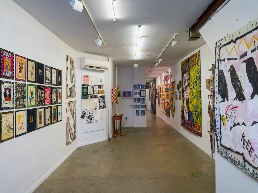 Gallery interior with various colorful paintings and artworks hanging on white walls, bordered by a wooden floor and track lighting.