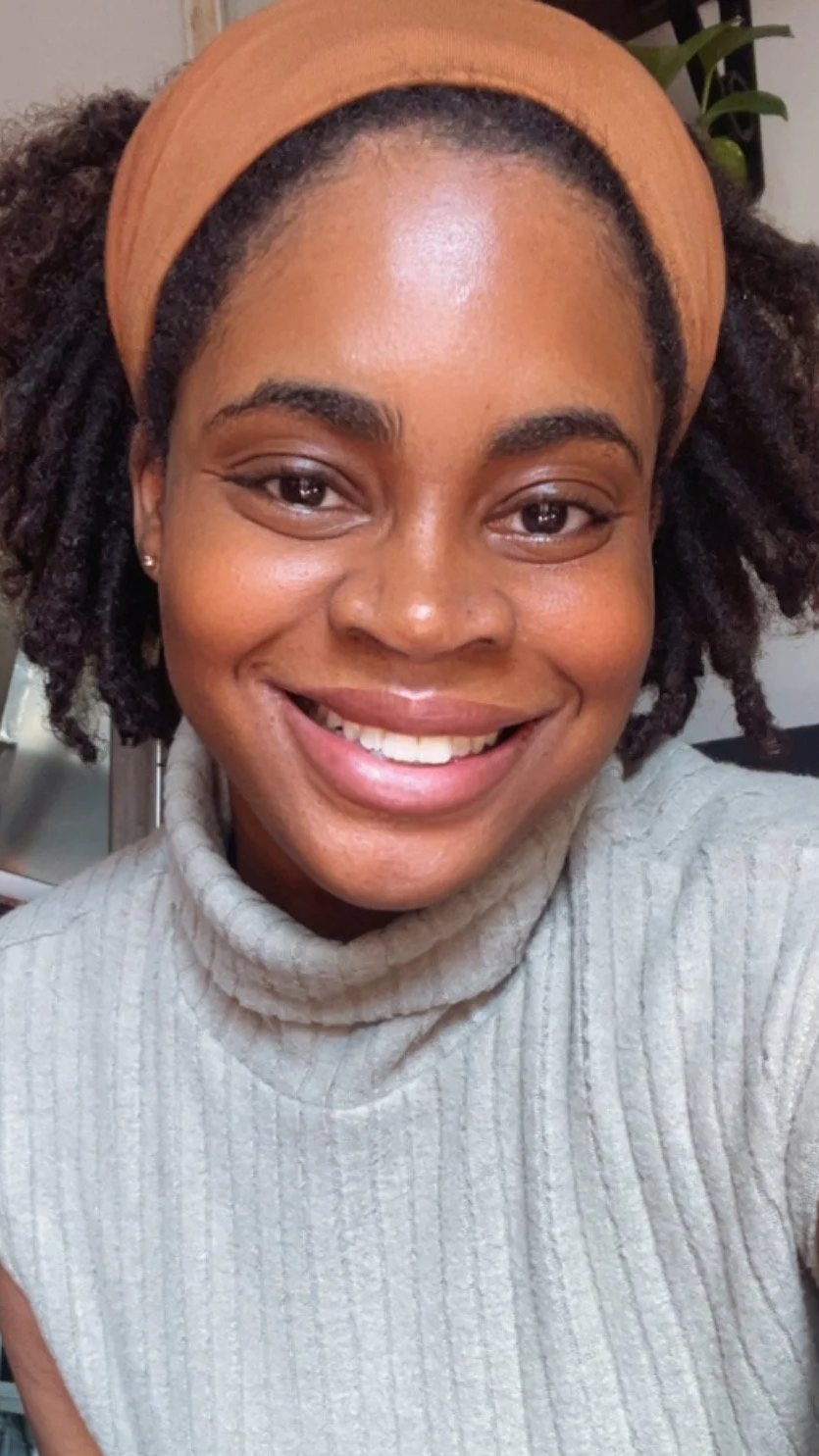 Close-up of a smiling woman with curly hair, wearing a beige headband and a gray turtleneck sweater.
