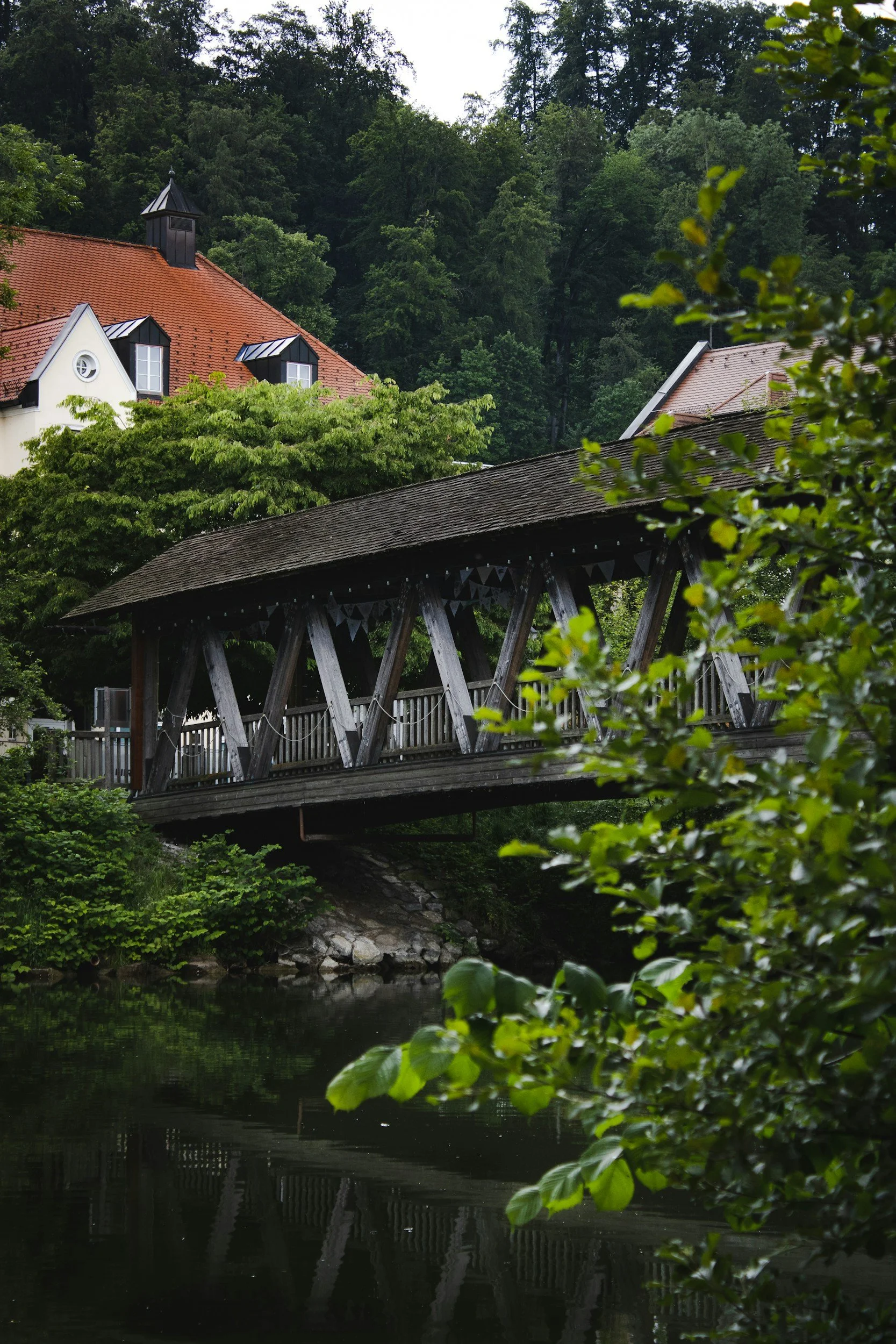 Ein Holzbrücke mit Dach spannt sich über einen ruhigen Fluss, umgeben von grünen Bäumen und Häusern mit roten Dächern im Hintergrund, in einer bewaldeten Hügellandschaft.