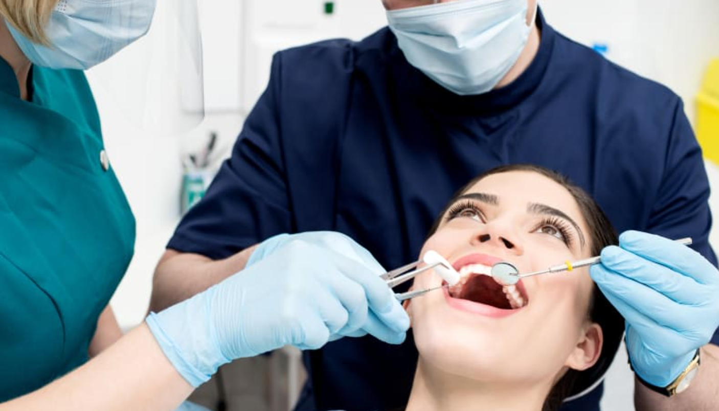 A young woman is at the dentist's office with a dentist and dental assistant examining her open mouth. The dentist is using dental tools, and all are wearing face masks and gloves for safety.