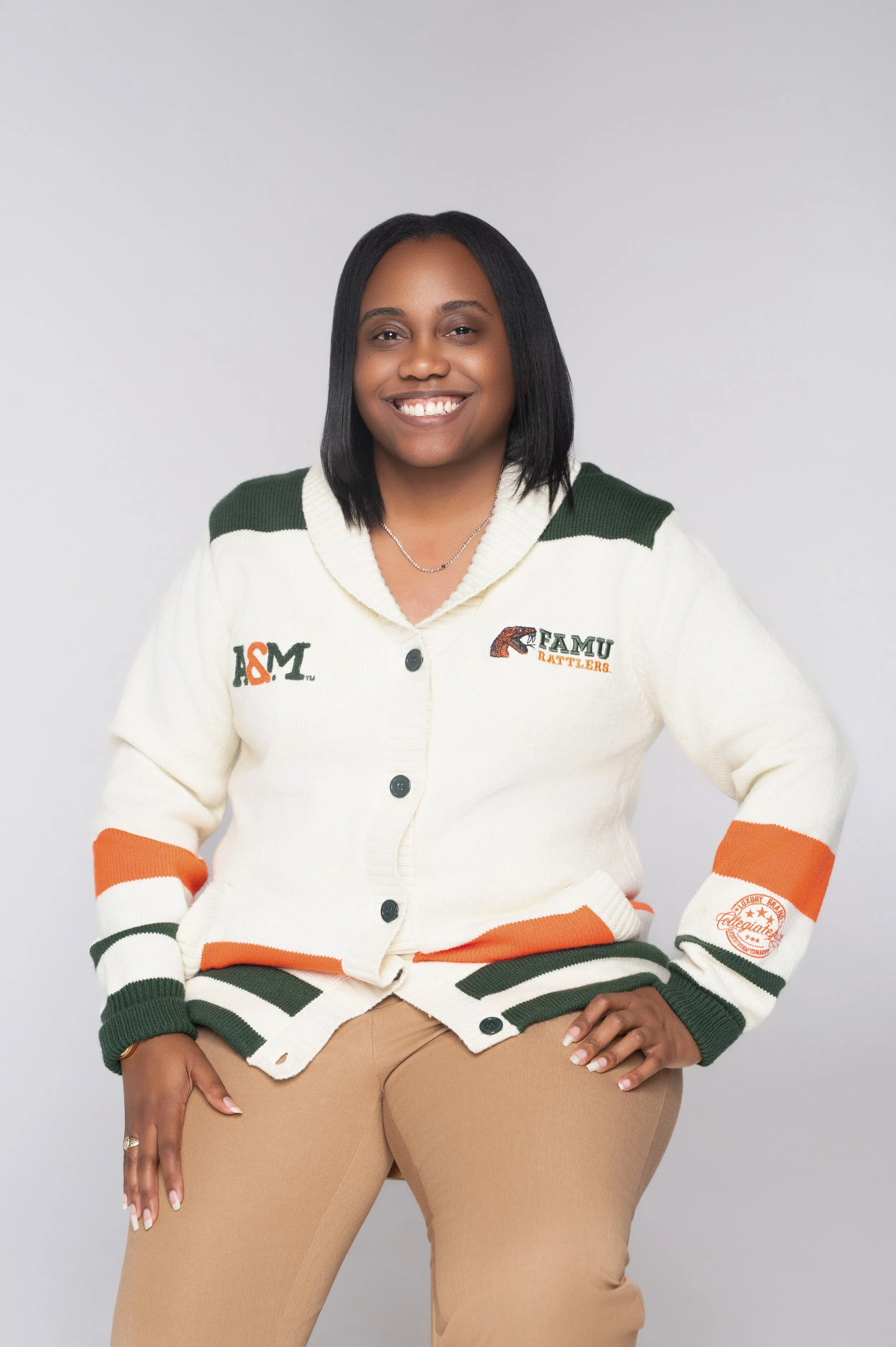A woman with black hair, wearing a cream-colored cardigan with green, orange, and black accents, and khaki pants, posing against a plain light gray background, smiling at the camera.