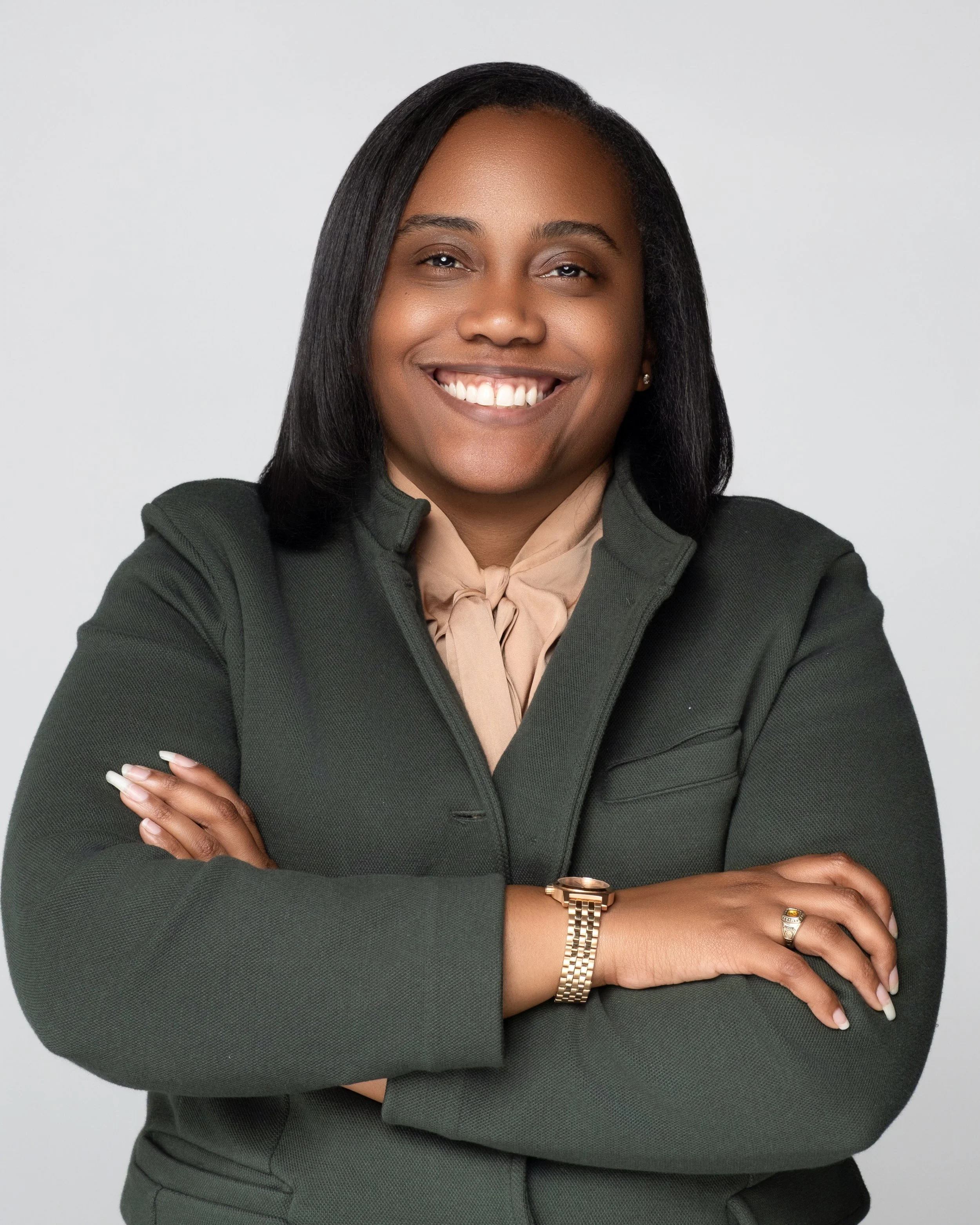 Woman with dark hair smiling, arms crossed, wearing a dark blazer, beige blouse, gold watch, and wedding ring