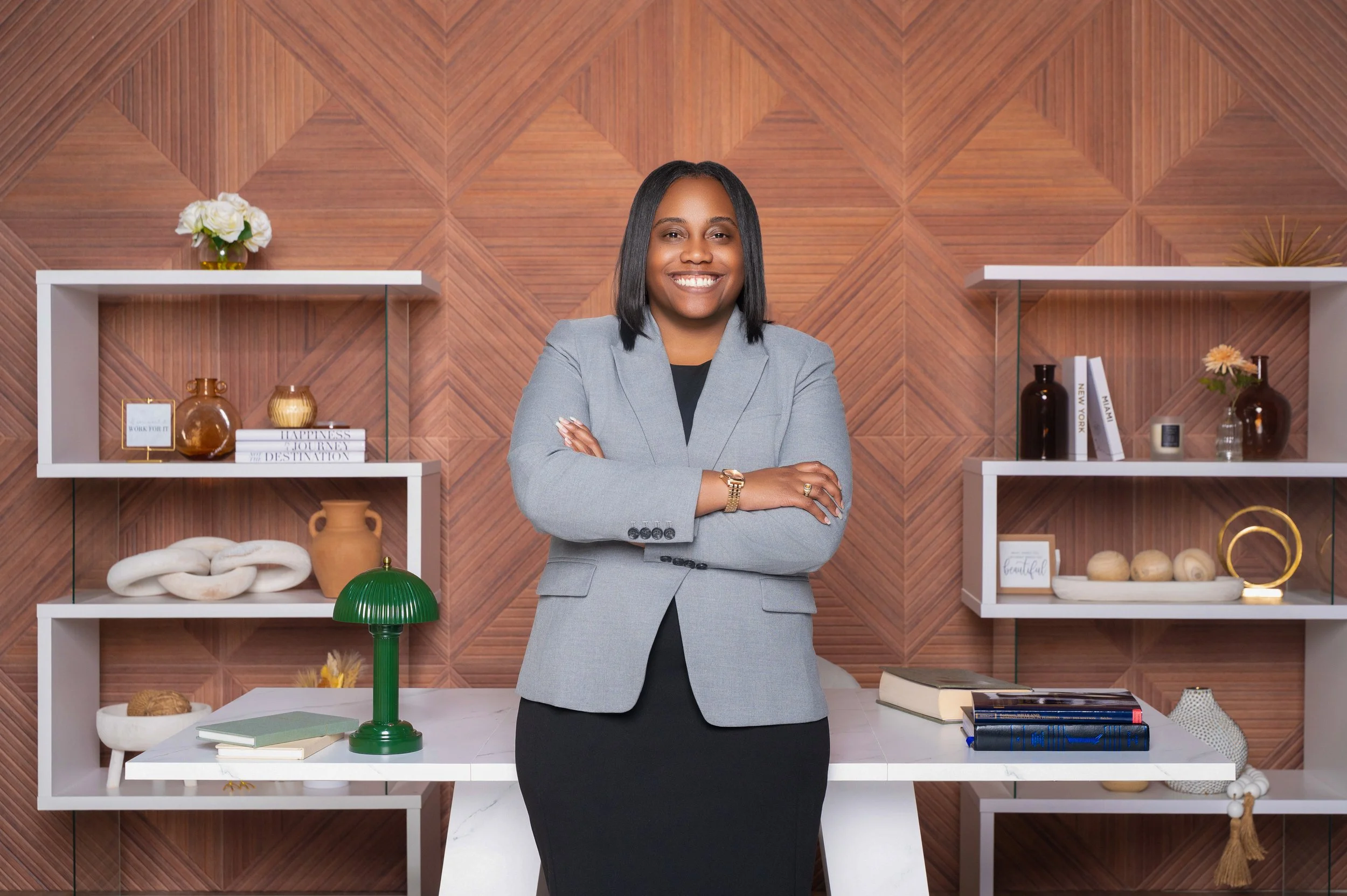 A woman with shoulder-length black hair, wearing a gray blazer and black dress, smiling with arms crossed, standing in front of a modern decorated wall with white shelves holding vases, books, and decor items.
