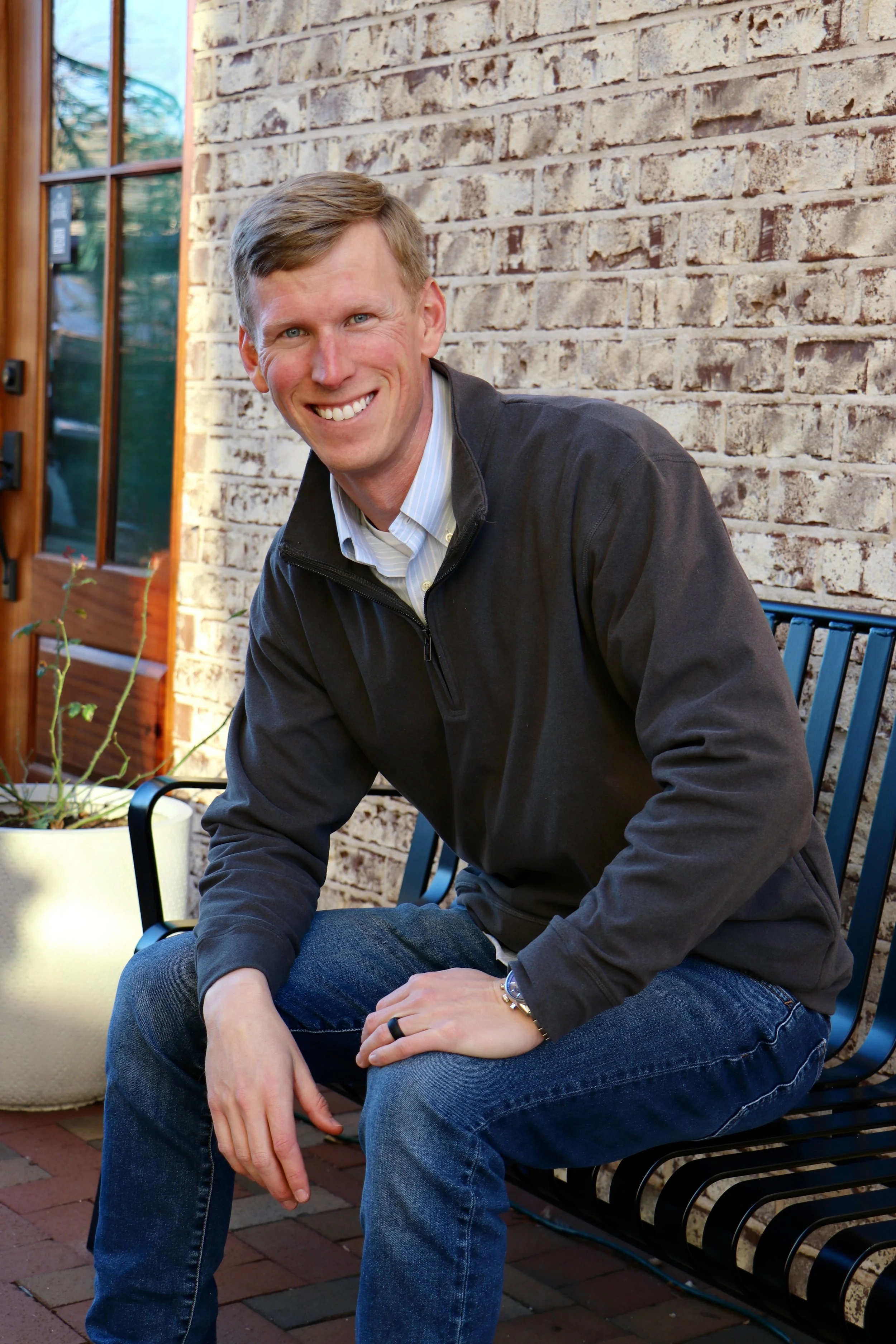 A smiling man with light brown hair, wearing a dark jacket and jeans, sitting on a black metal garden bench outside next to a potted plant and a brick wall.