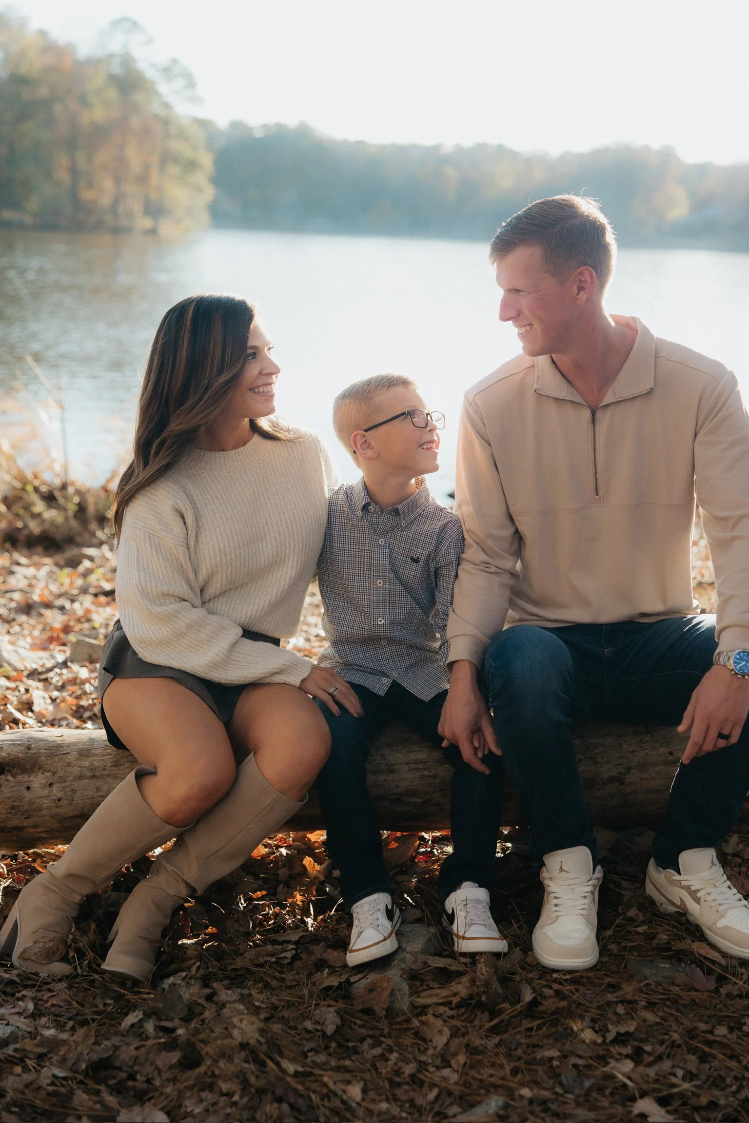 A family of three sitting on a log near a lake, smiling and looking at each other, with autumn leaves on the ground.