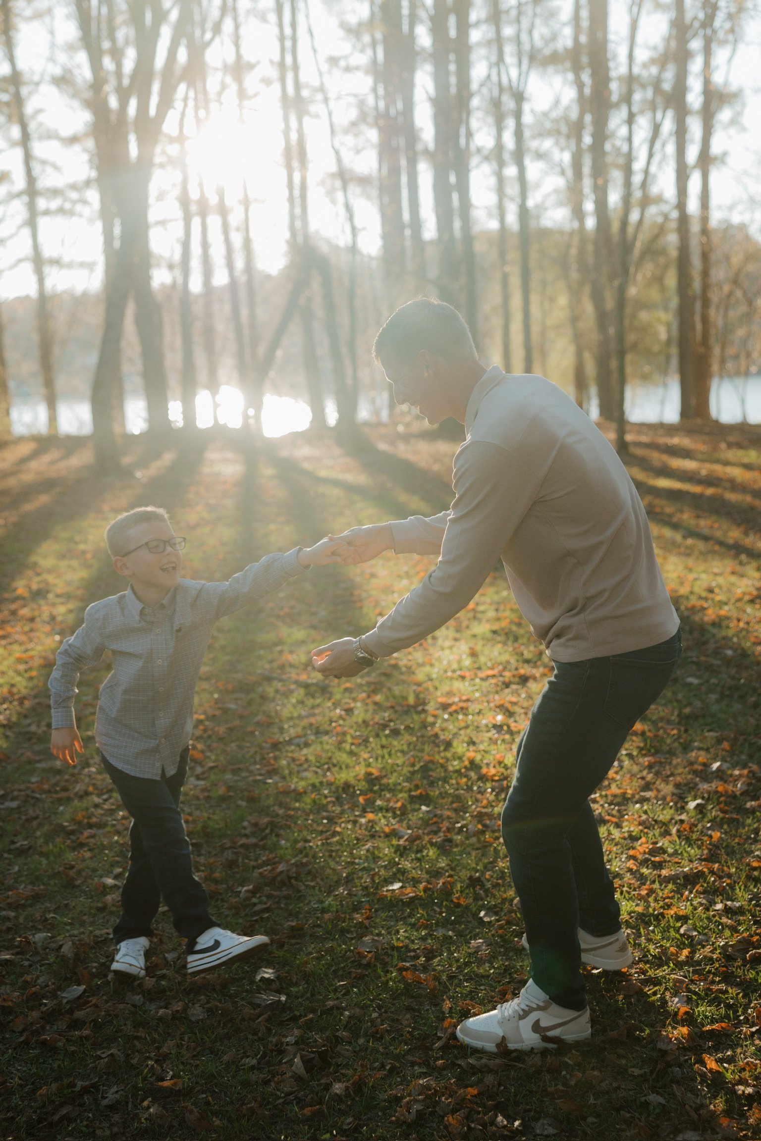 A man and a boy playing together in a wooded park during fall, with sunlight filtering through trees, as the man hands something to the boy, who is smiling.