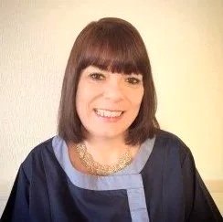 A woman with shoulder-length dark hair and bangs smiling, wearing a dark top with a graphic design and a sparkly necklace.