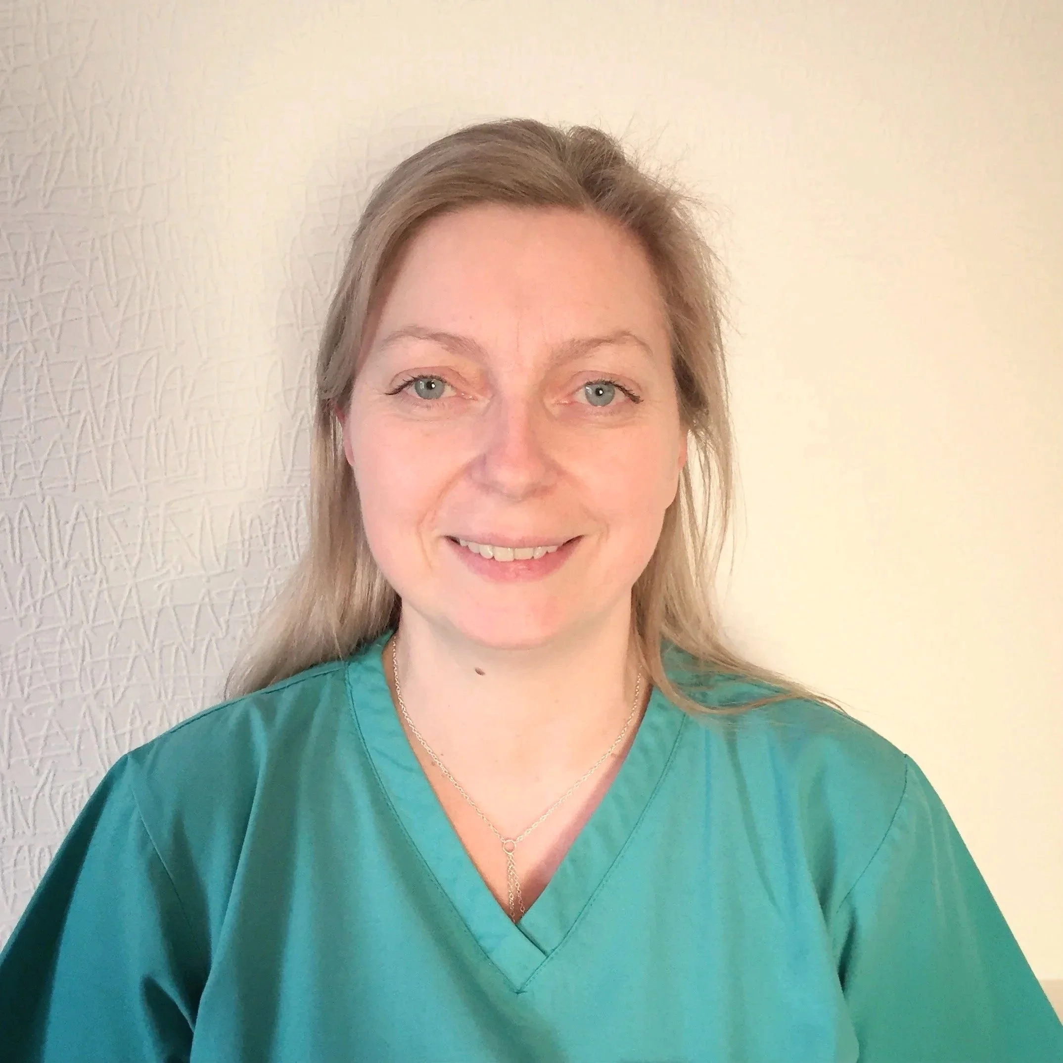 A woman with blonde hair wearing teal scrubs, smiling, standing against a textured light-colored wall.