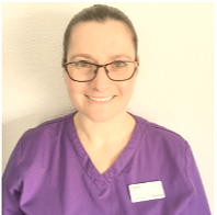 Smiling woman wearing glasses and a purple scrub top with a name badge, standing against a light-colored wall.