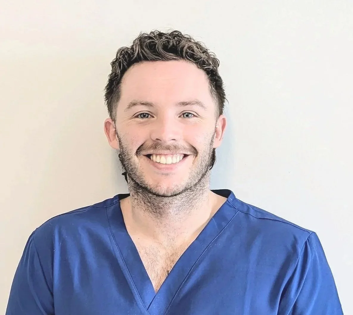 A smiling man with dark, curly hair and light skin, wearing blue scrubs, standing against a plain white wall.