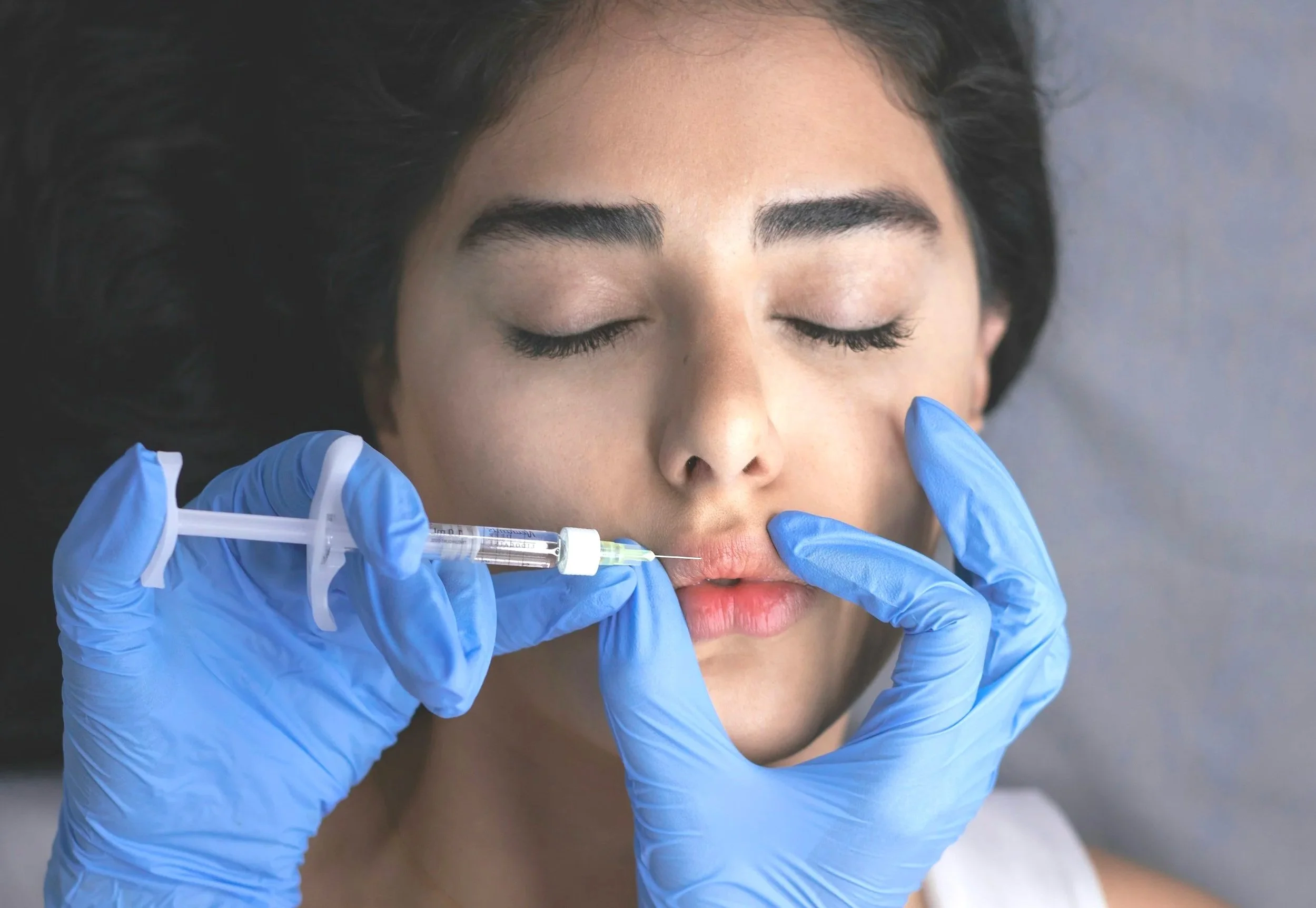 A woman with closed eyes lying down, receiving a cosmetic lip injection from a medical professional wearing blue gloves.