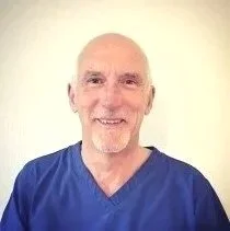 Portrait of an elderly man with a bald head and a white goatee, wearing a blue shirt, smiling against a plain light-colored background.