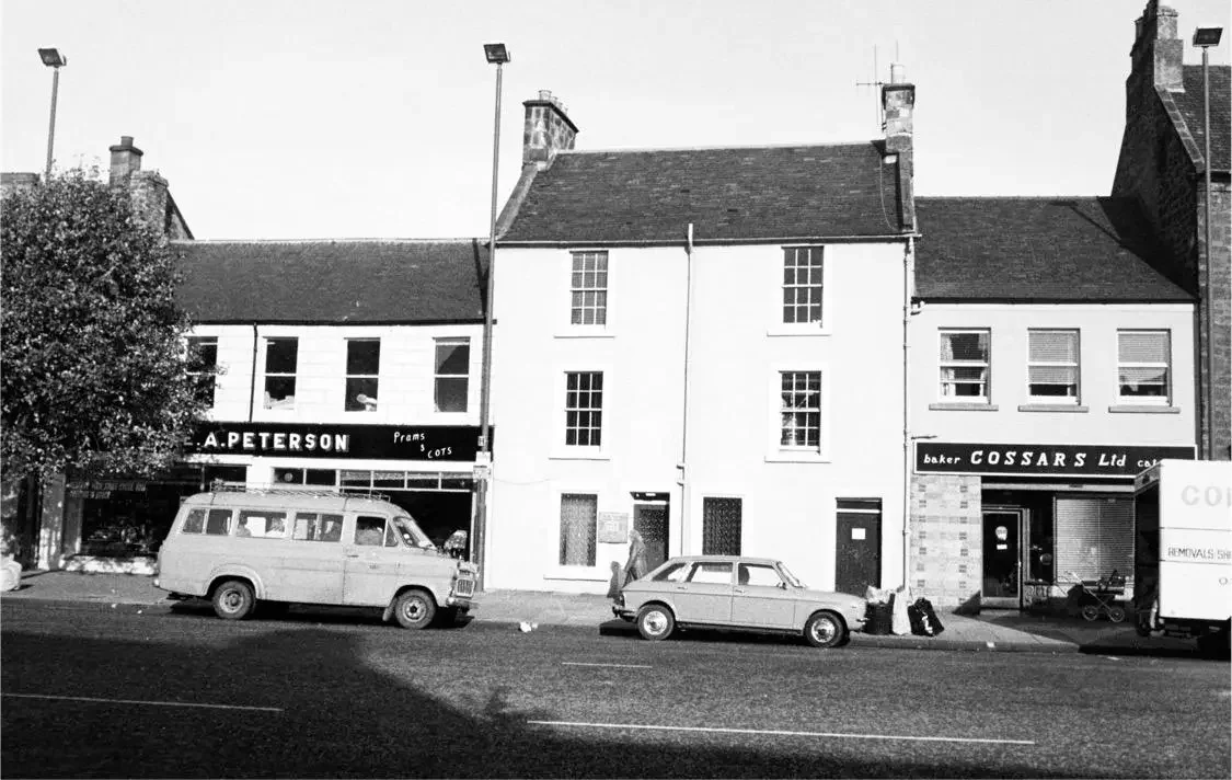 Black and white photo of a street with parked cars in front of row of buildings, including a shop named E. A. Peterson and another named Cossars Ltd.