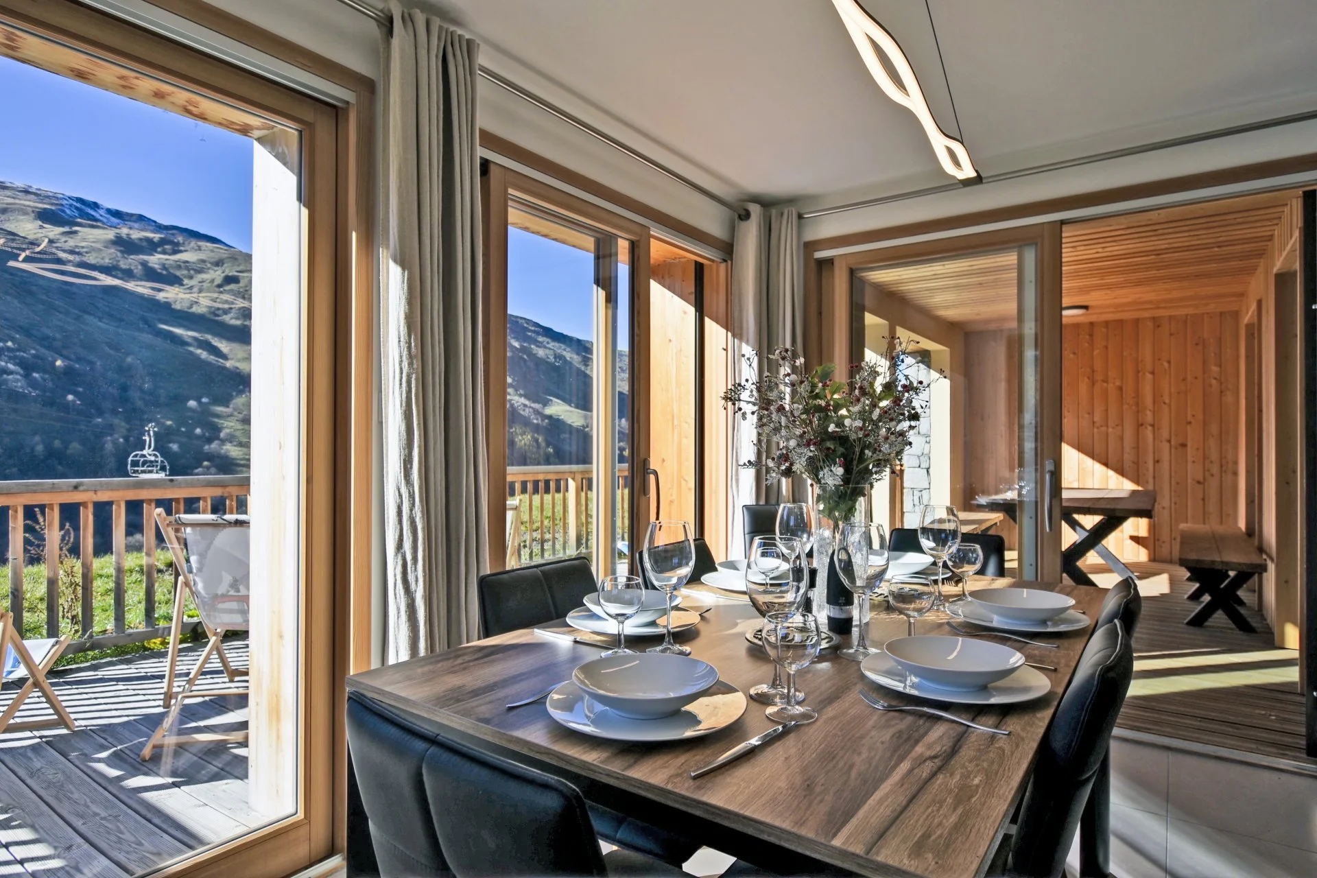 Salle à manger avec une table en bois, dressée avec des assiettes, verres et une grande vase de fleurs. La pièce est lumineuse avec de grandes fenêtres donnant sur un balcon avec vue sur des montagnes.