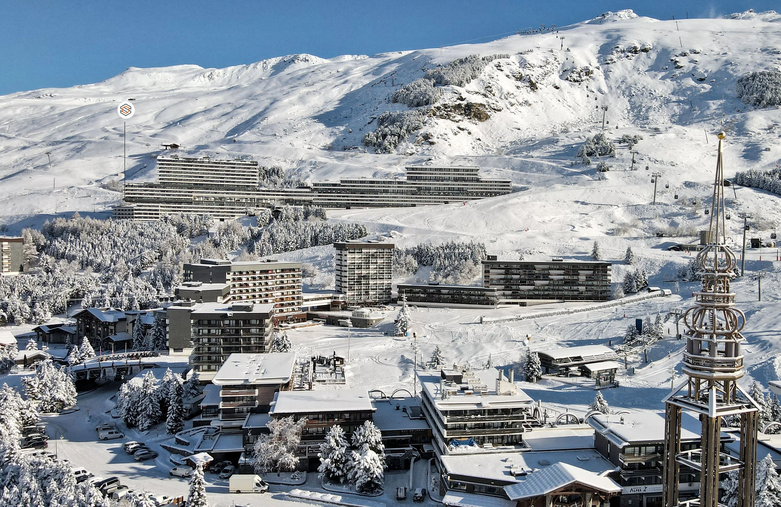 Vue d'une station de ski avec des bâtiments modernes recouverts de neige, une pente enneigée avec des remontées mécaniques, et un ciel clair.