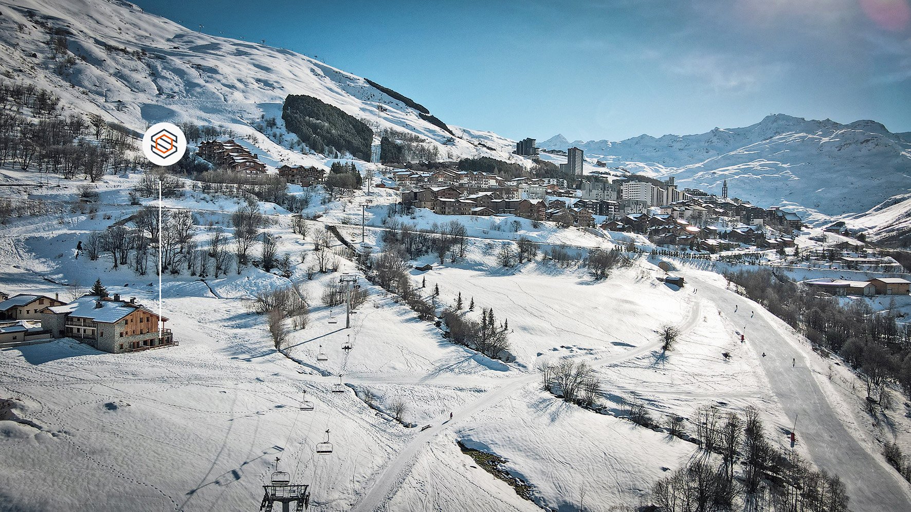 Paysage de montagne enneigée avec des bâtiments, des arbres, des pistes ski et des remontées mécaniques