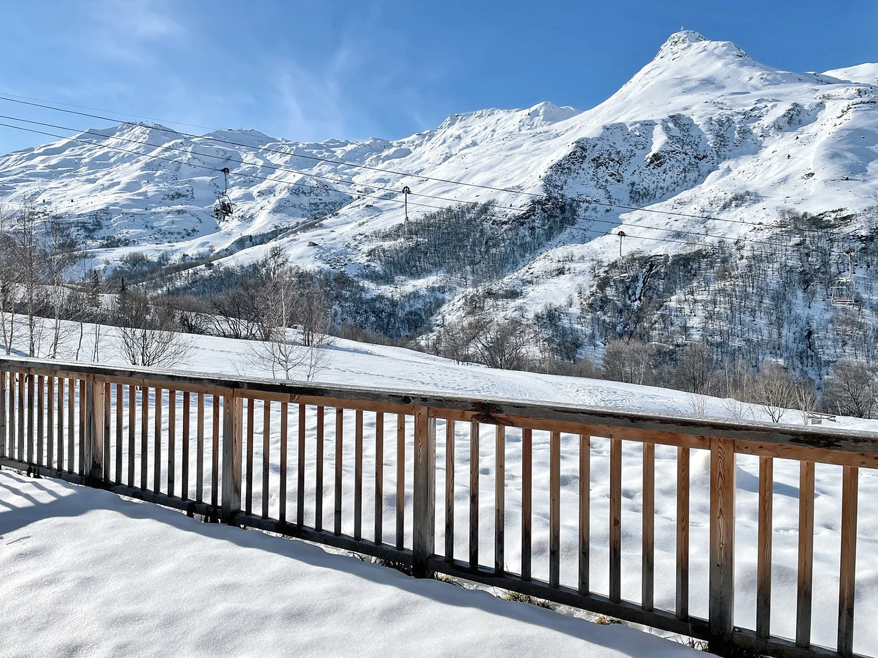 Vue d'une montagne enneigée avec un ciel bleu clair vue depuis une terrasse en bois enneigée, avec des télécabines en suspension dans l'air.