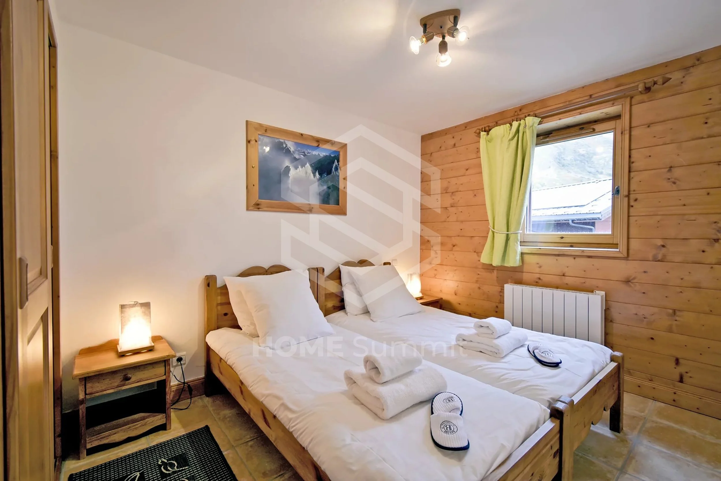 Chambre avec deux lits en bois, une fenêtre avec rideau vert, un tableau représentant des montagnes, lampes de lecture et serviettes pliées sur les lits.