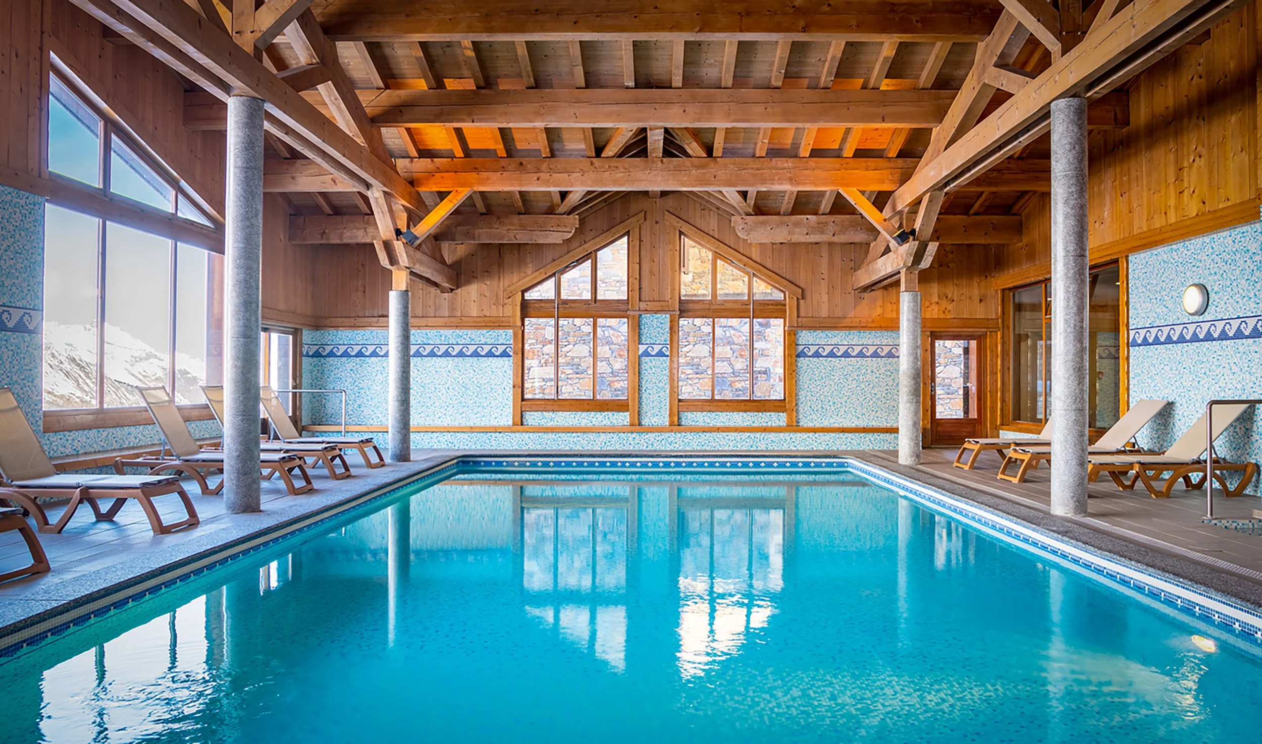 Piscine intérieure dans une cabane de montagne en bois, avec chaises longues et grande fenêtre offrant une vue sur la montagne enneigée.