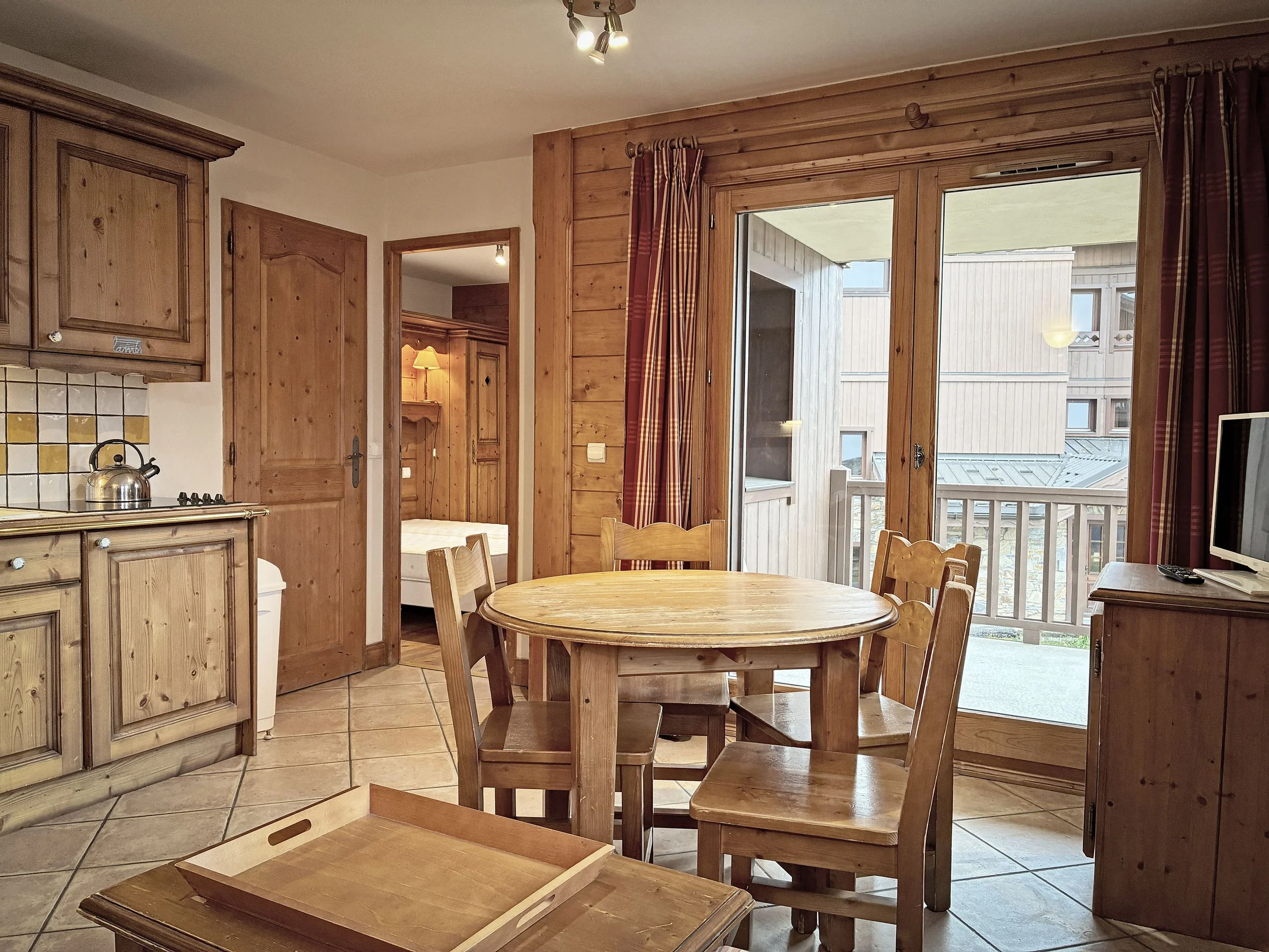 Intérieur d'une cuisine avec mobilier en bois, table ronde et chaises, accès à un balcon par une porte-fenêtre, et une télévision sur un meuble en bois.